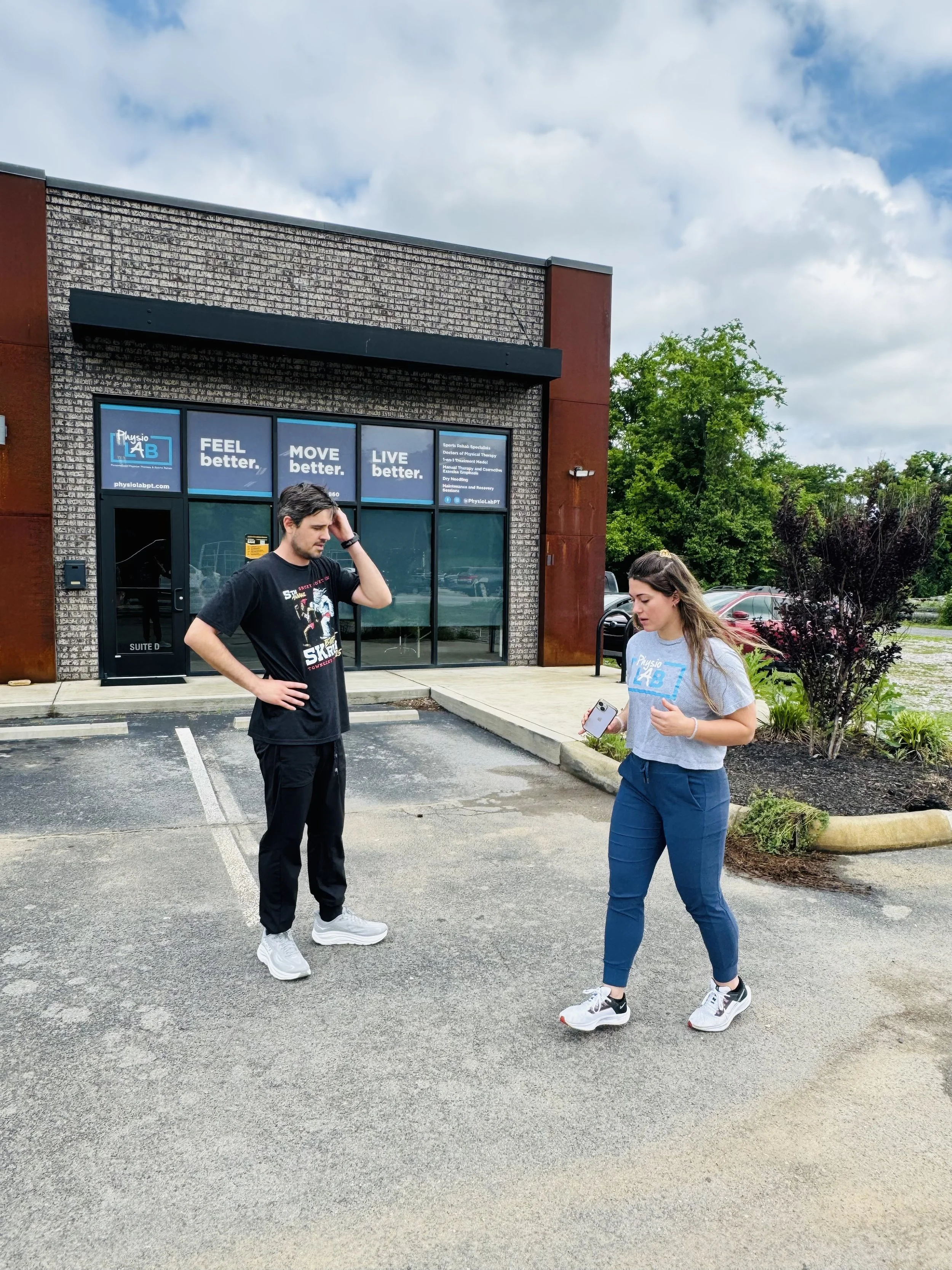 A man and a woman standing in a parking lot outside a fitness center, with the woman looking at her phone and the man touching his head. The fitness center has signage that says 'Feel better. Move better. Live better.' and features a brick facade and large glass windows.