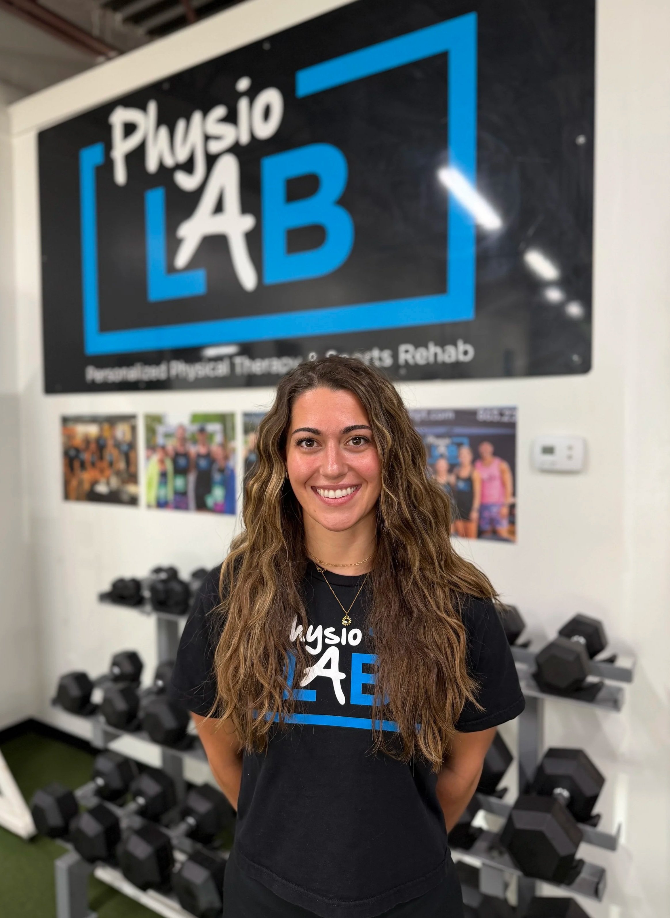 A woman with long, curly brown hair smiling in a gym, standing in front of a sign that reads 'Physio LAB' and a wall of black dumbbells.
