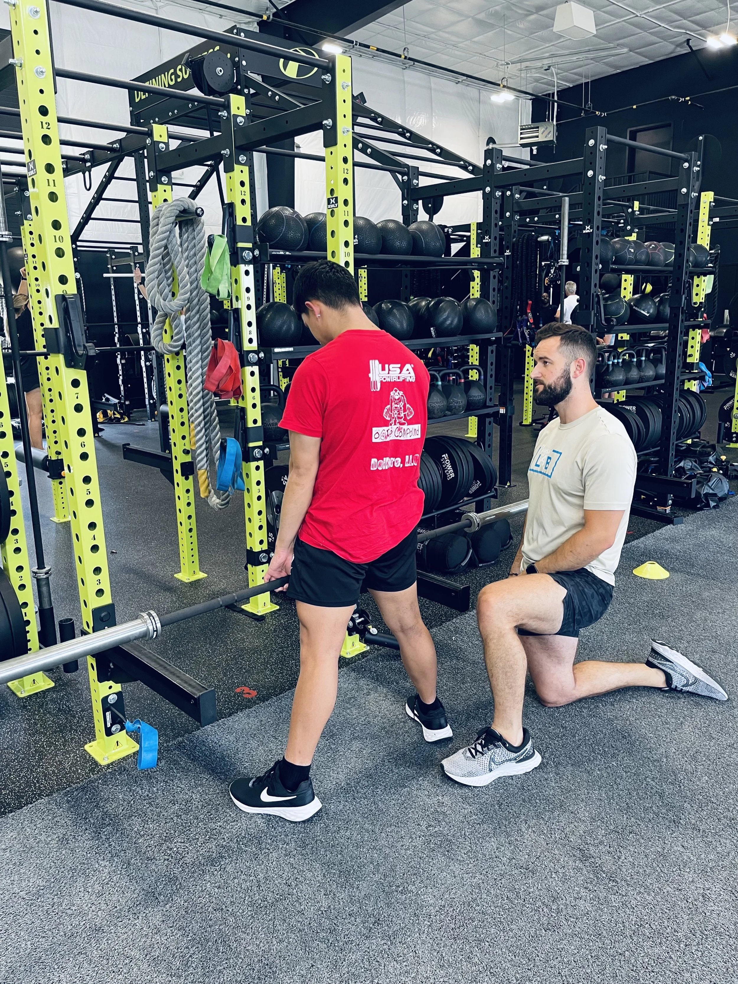 Two men in a gym, one kneeling and the other standing, preparing for a lift near a black and yellow power rack with weights and equipment.