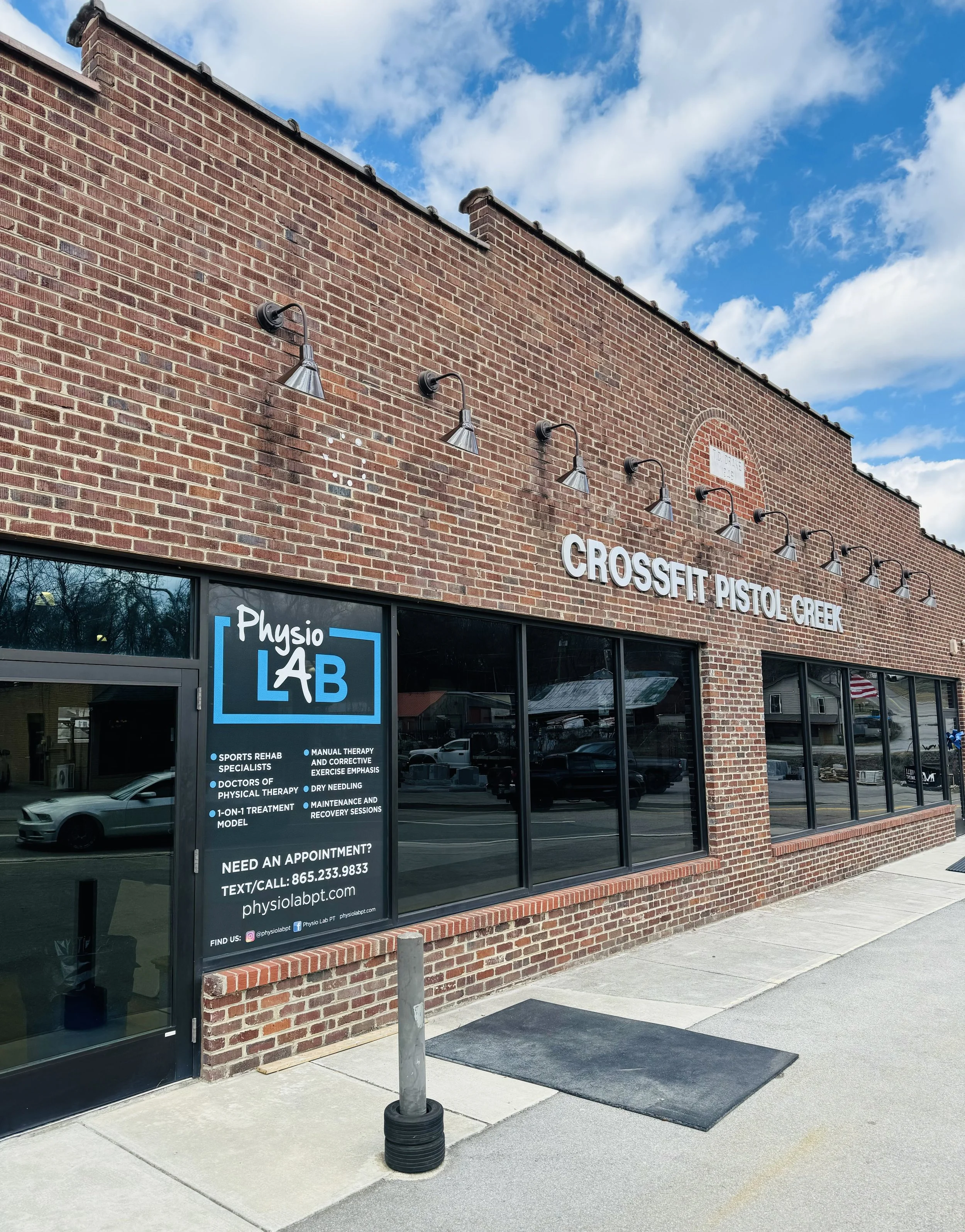 A brick building with a sign indicating it houses a Crossfit gym and a physical therapy clinic. The sign features details about services, appointments, and contact information, with a storefront window reflecting trees and parked cars outside.