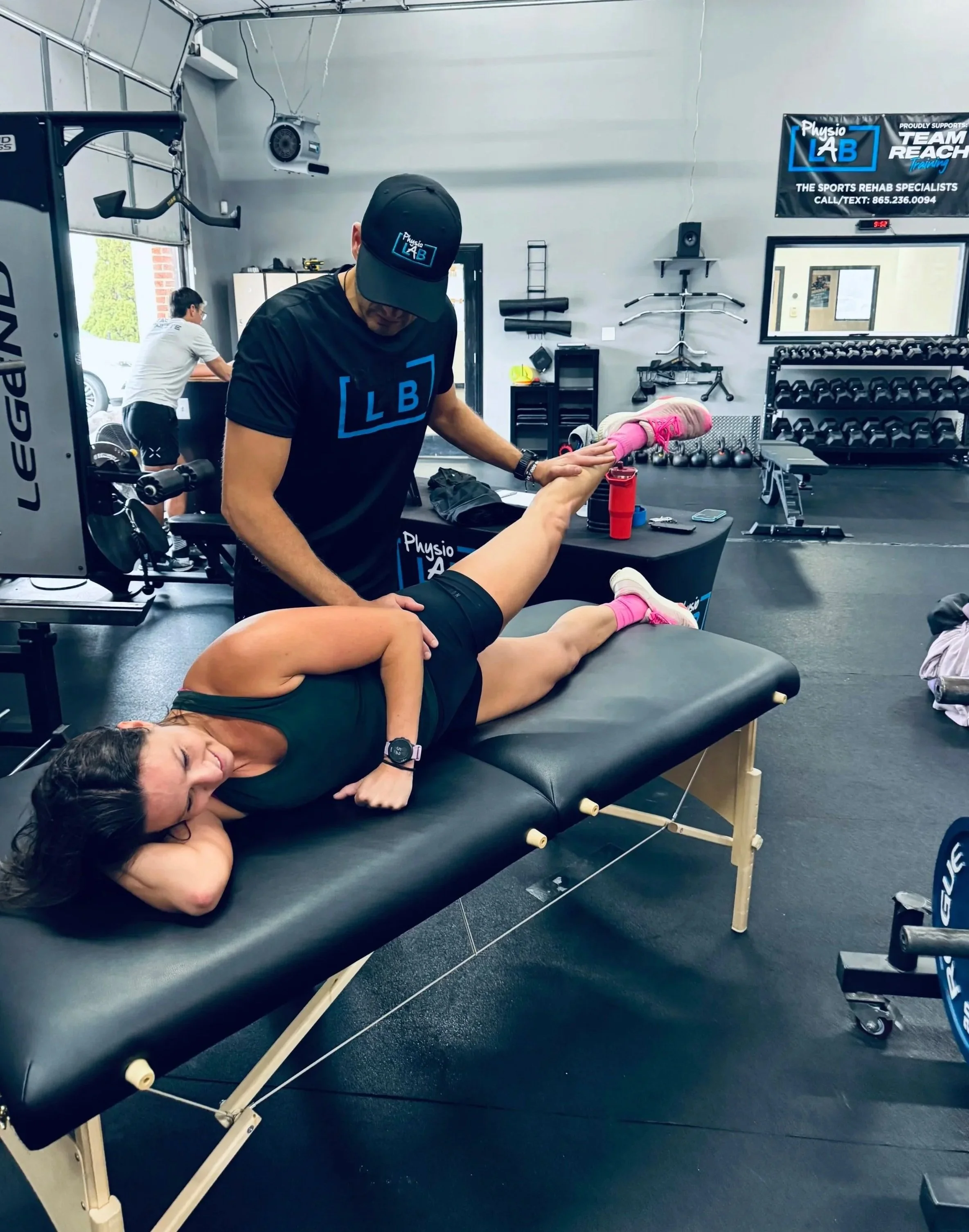 A woman lying face down on a massage table wearing a black tank top, with her eyes closed and smiling, undergoing a physical therapy session. A physical therapist in a black shirt and cap with 'Physio LAB' logo is assisting her. In the background, gym equipment including kettlebells, barbells, and a wall-mounted rack of weights are visible.