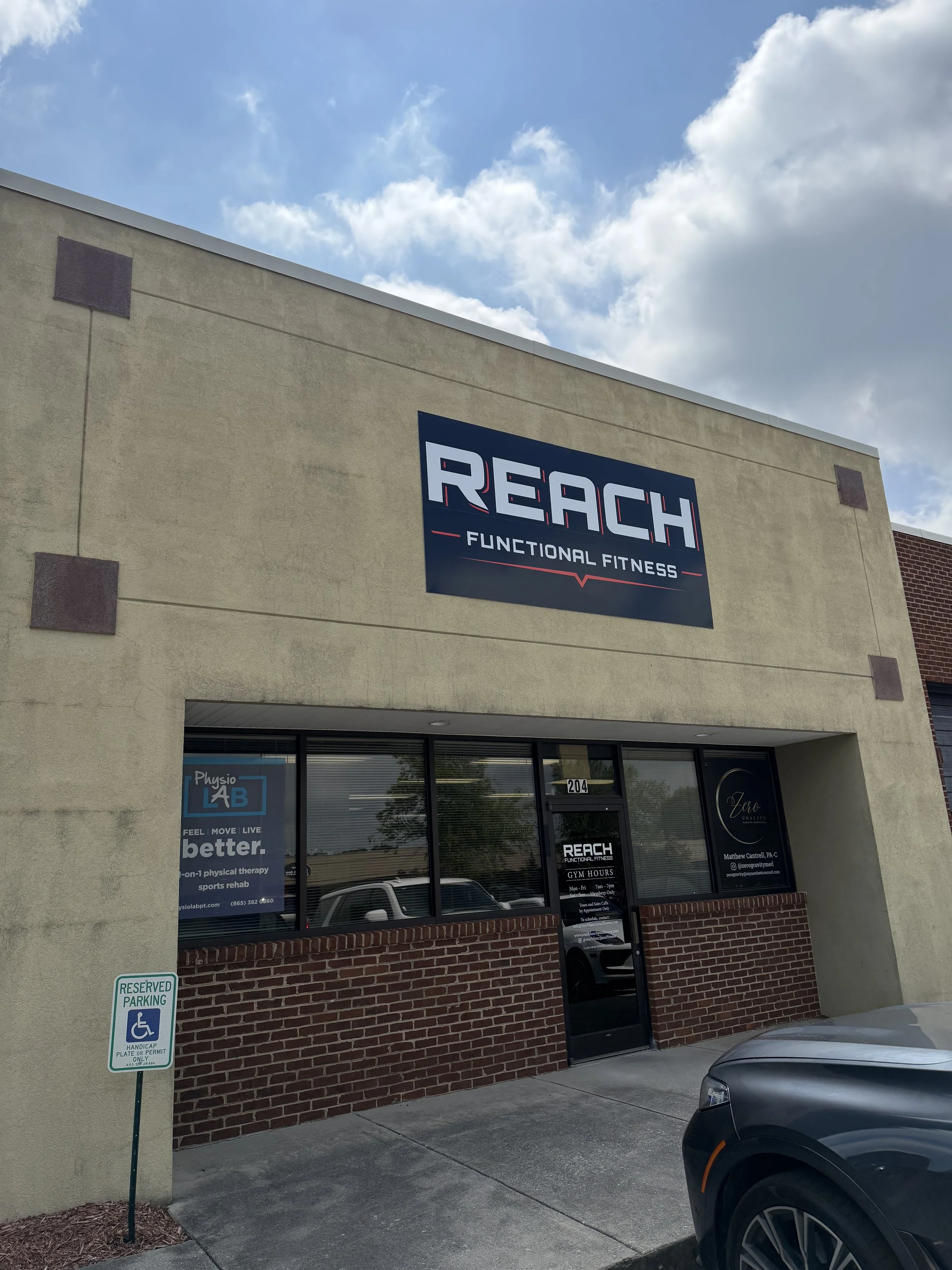 The image shows the front of a building with a sign that reads 'REACH FUNCTIONAL FITNESS.' There are windows below the sign, some of which display advertisements for physical therapy and health services. A reserved parking sign is visible on the left, and nearby cars are parked in front of the building. The sky above is partly cloudy.