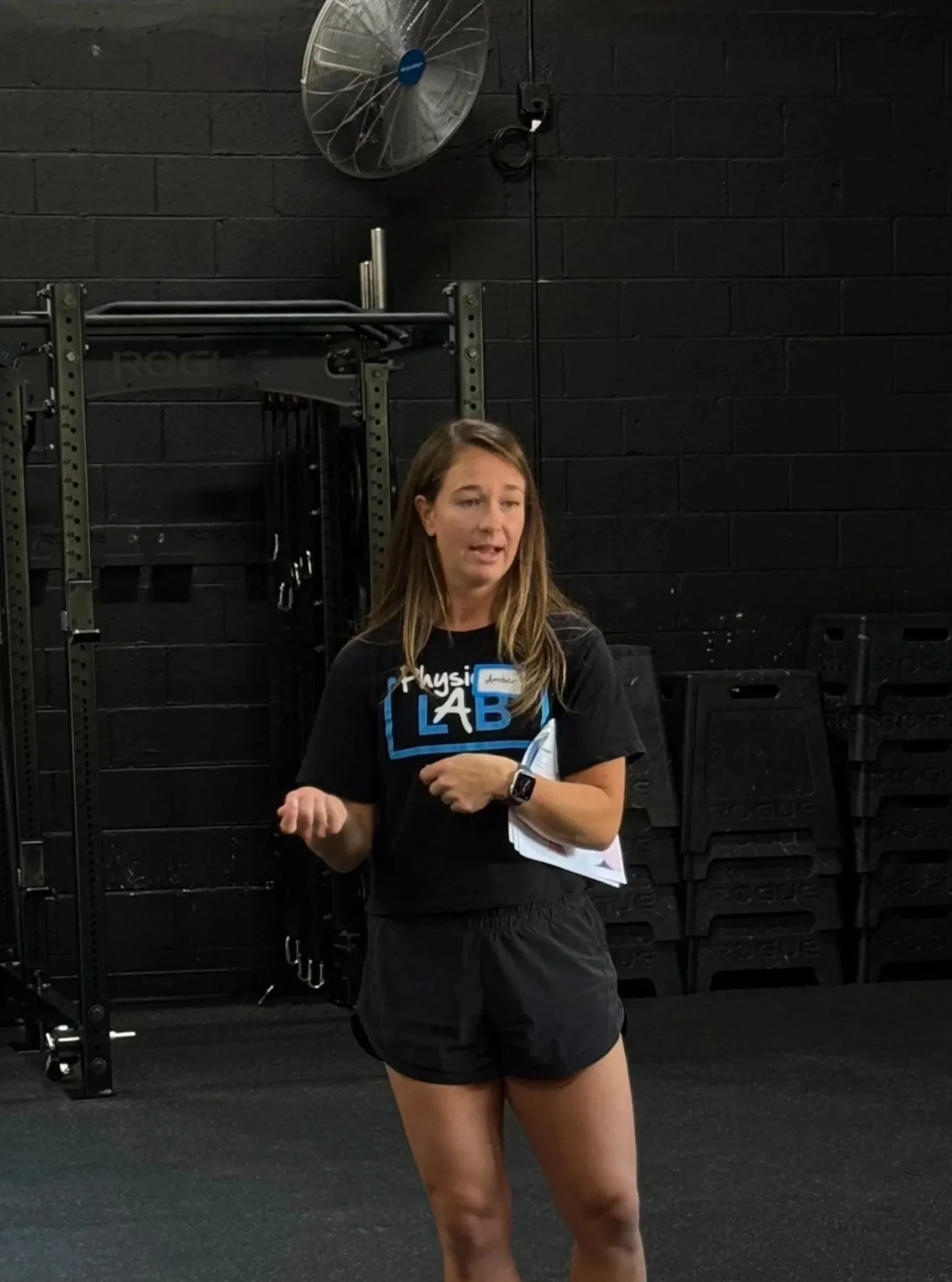 A woman with long hair standing in a gym, wearing a black t-shirt with the word 'LAB' on it, black shorts, and a fitness watch, holding a notebook, in front of a black brick wall, with a large industrial fan and gym equipment in the background.