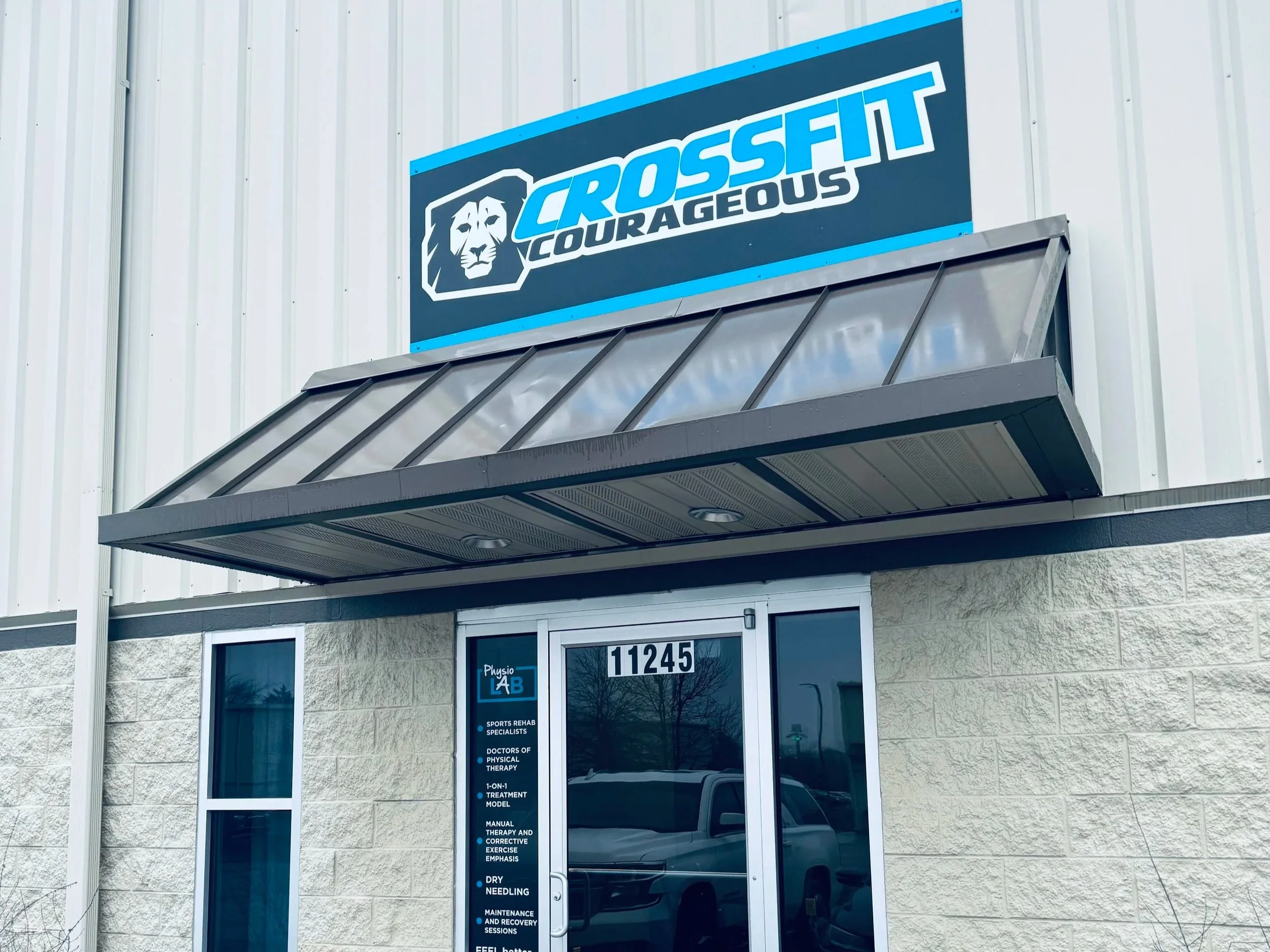 Sign for CrossFit Courageous gym mounted on the exterior wall of a building, with a metal awning below and windows displaying text for services offered.