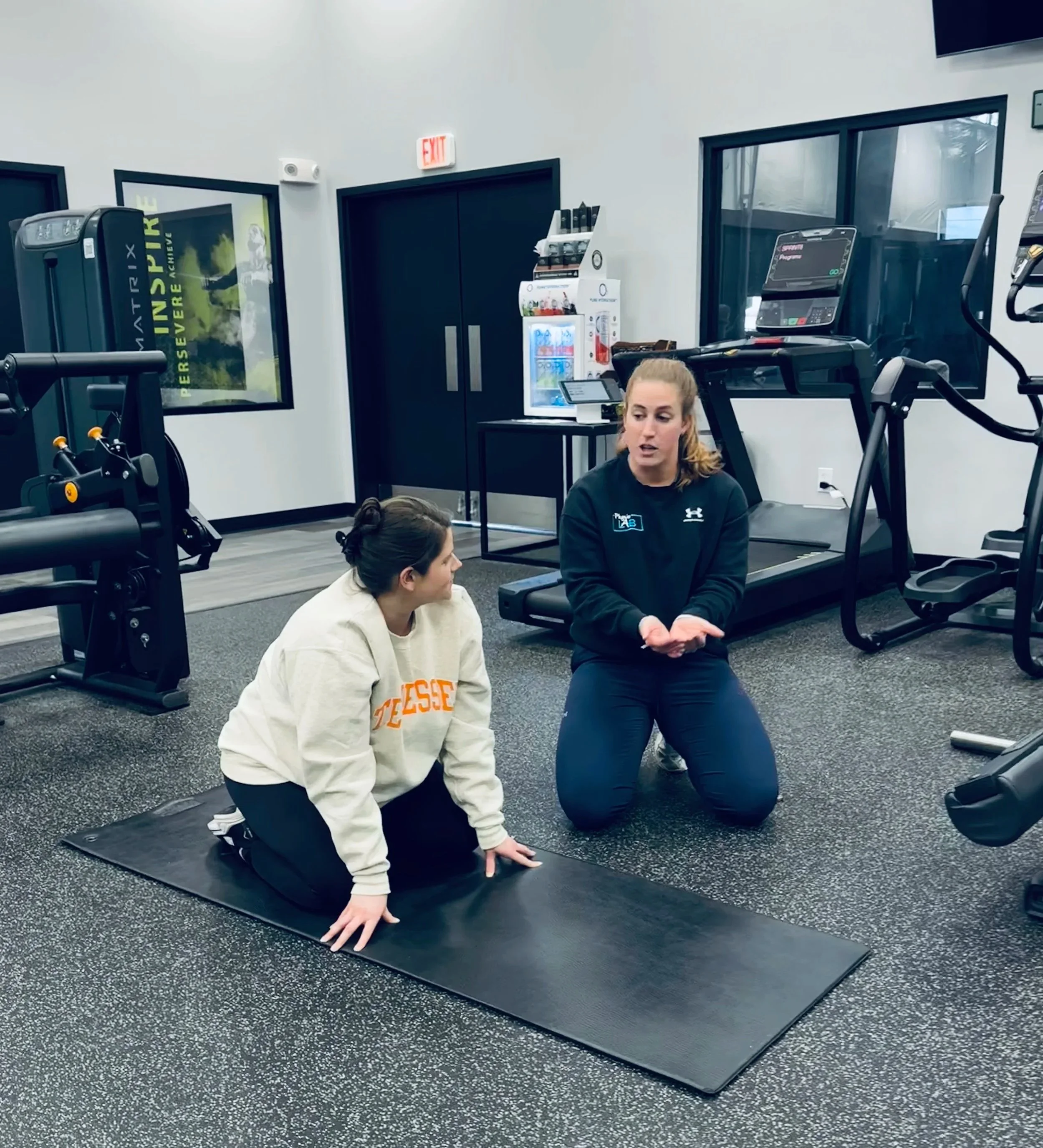 A woman in a black shirt is demonstrating an exercise to a woman in a white hoodie on a grey exercise mat in a gym. The gym has exercise equipment, a vending machine, and a large window.