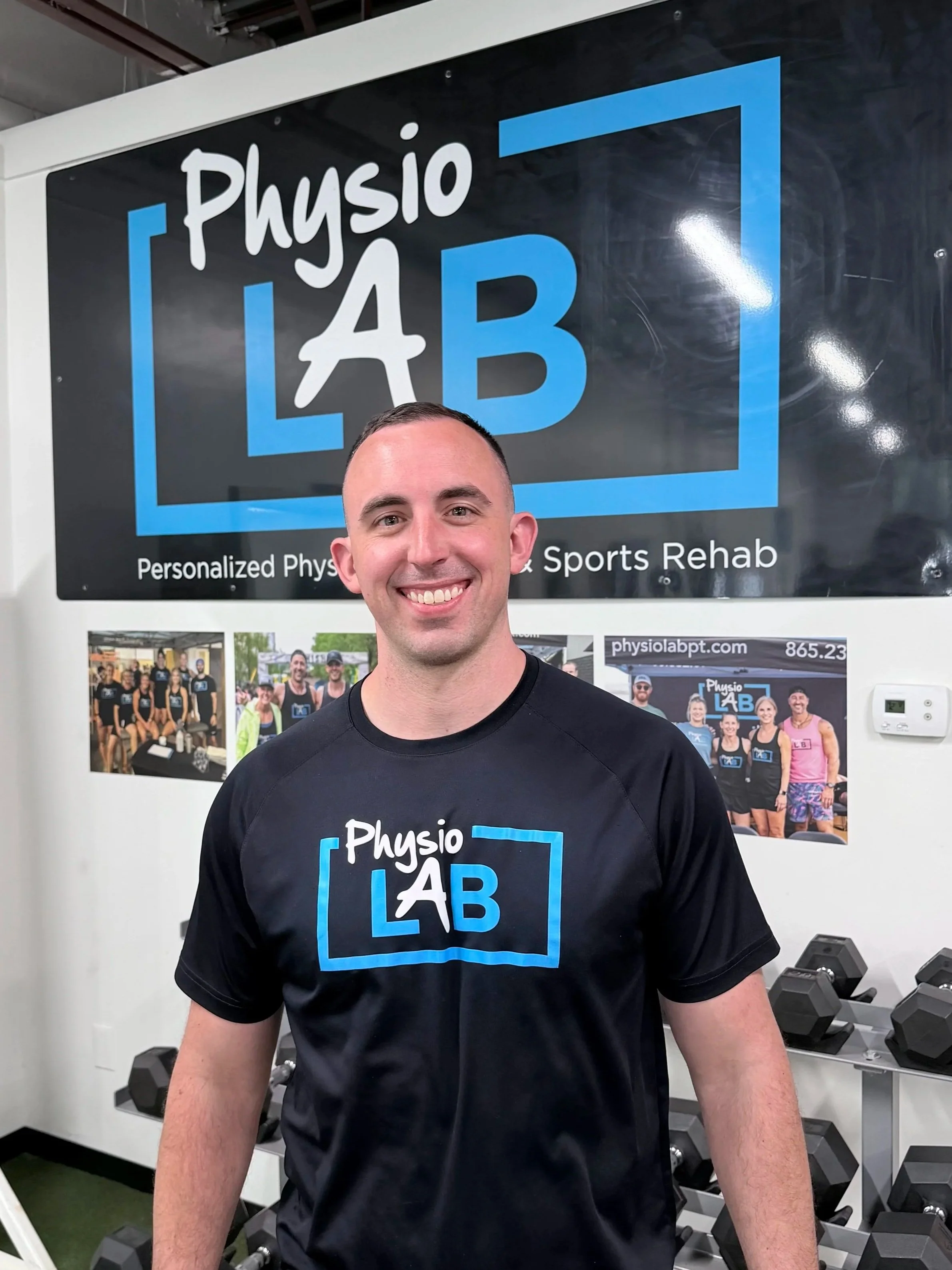 A smiling man in a black t-shirt standing in front of a large sign that reads 'Physio LAB' with a blue frame design, inside a gym or rehab center with dumbbells and photos on the wall.