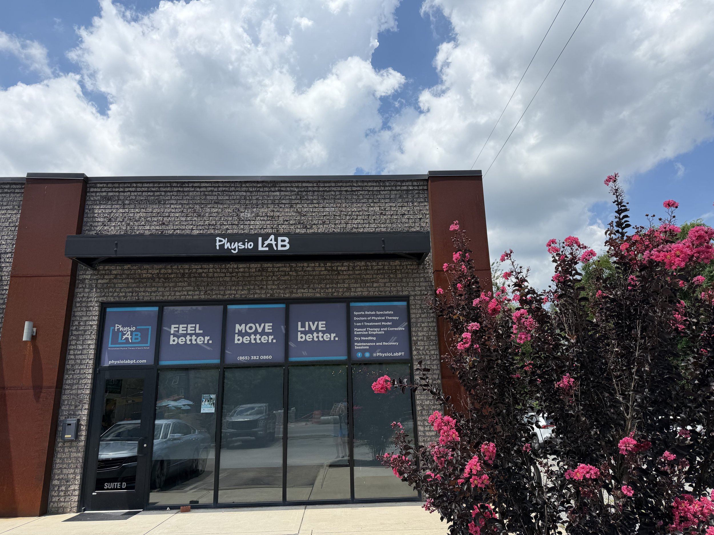 Exterior view of Physio LAB, a physical therapy and sports rehab clinic, showing the building's glass entrance door, signage, and pink flowering bushes in front under a partly cloudy sky.