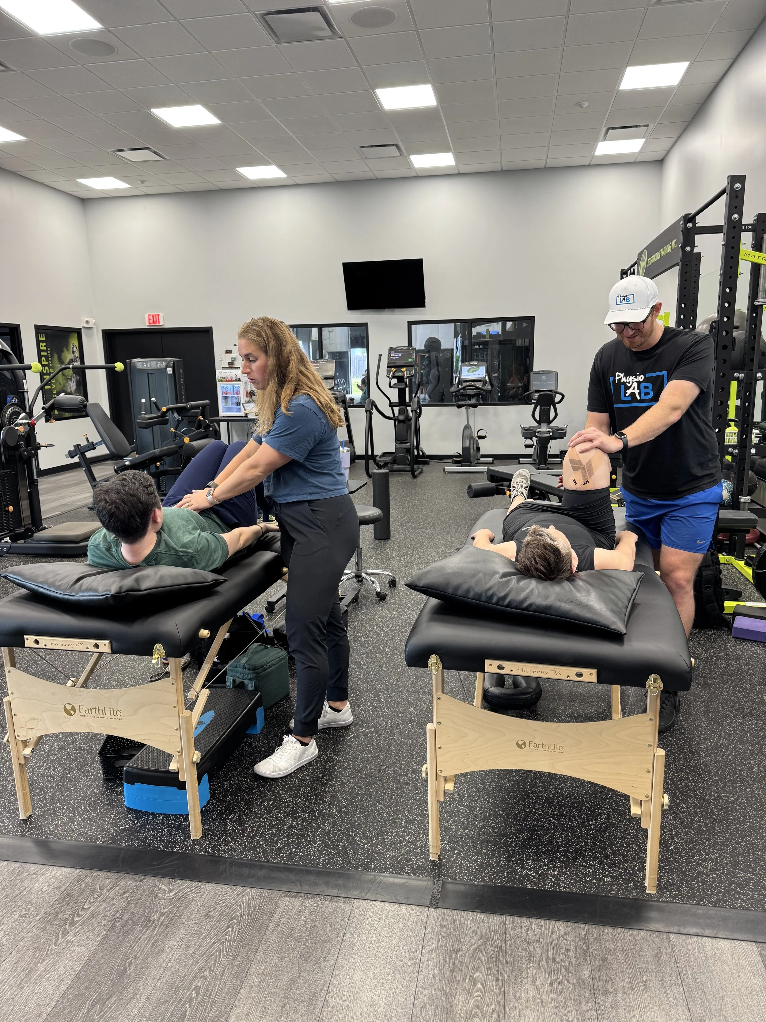 A physical therapy or chiropractic clinic with multiple people receiving treatment on adjustable massage tables, therapists providing care, and exercise equipment in the background. The room has a gray floor, white walls, and a ceiling with fluoresce