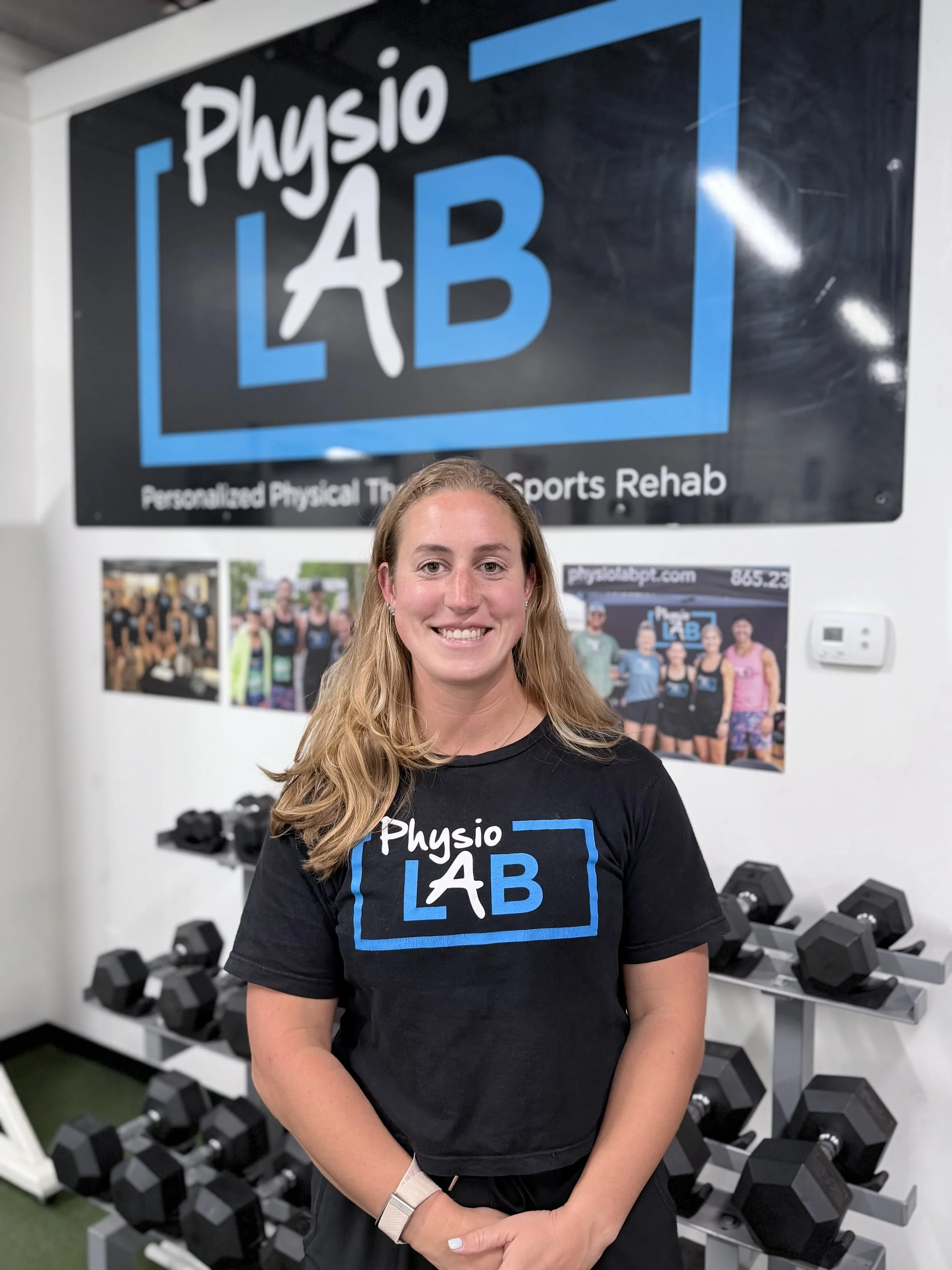 A woman with long blonde hair smiling at the camera, wearing a black shirt with a logo that reads "Physio LAB" in front of a wall with fitness posters and dumbbells in a gym or physical therapy setting.