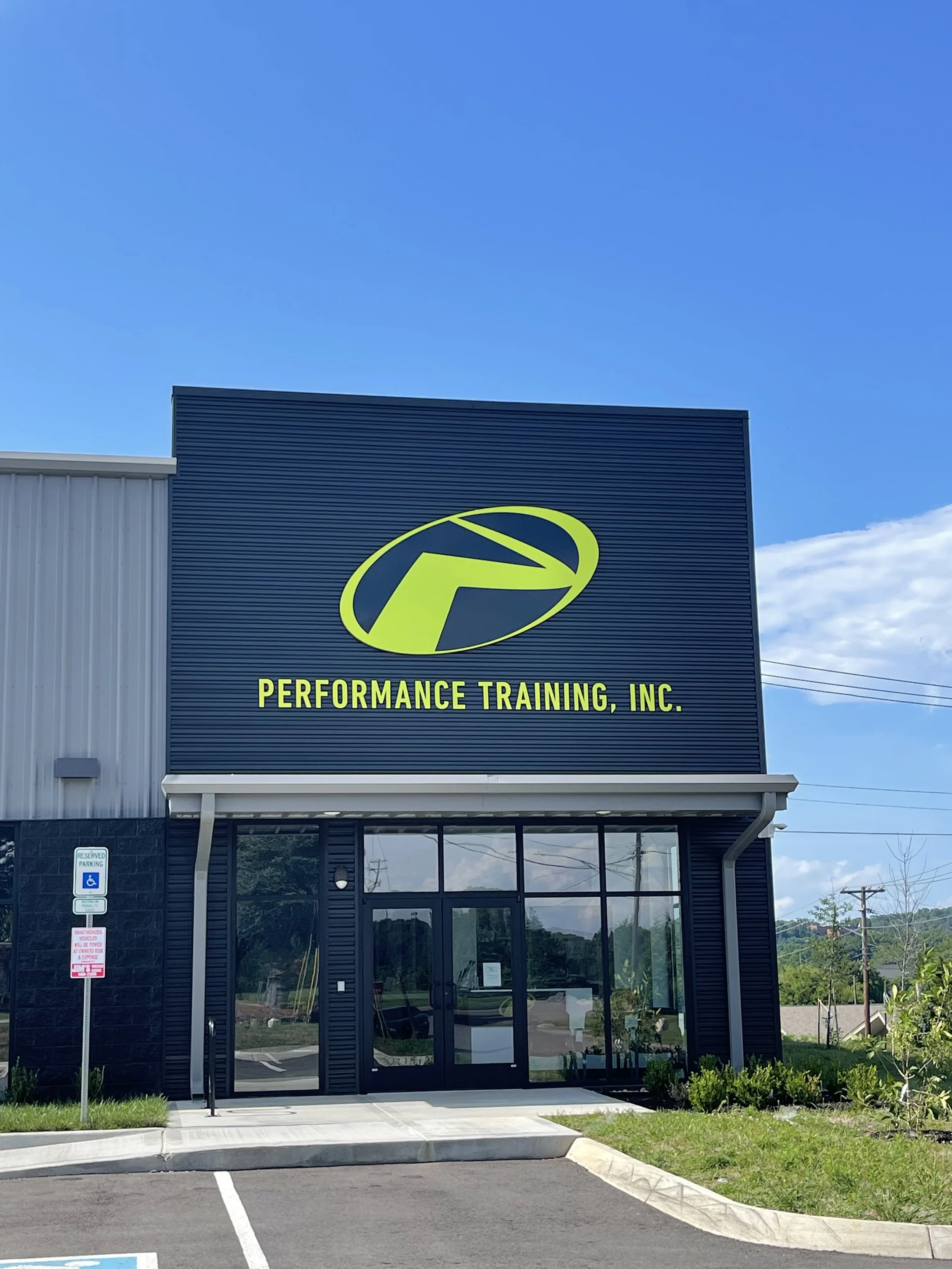 Front of a performance training gym with a large sign featuring a yellow and black logo and yellow text that reads 'Performance Training, Inc.' The building has a glass entrance door, a parking lot with a handicap parking sign, and some greenery in front.
