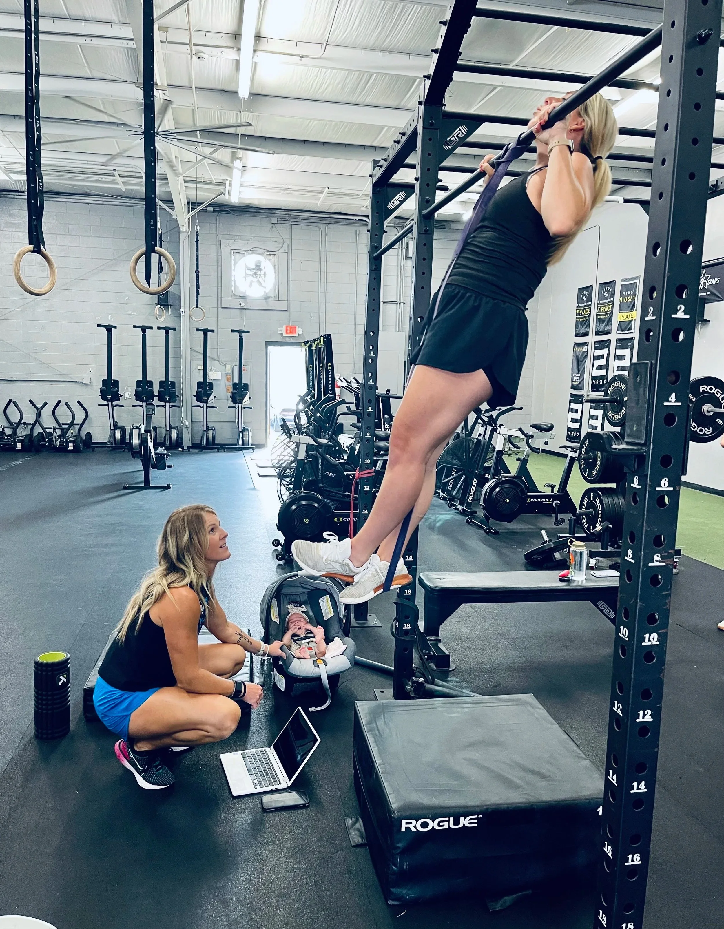 A woman is doing a pull-up exercise on a black power cage at a gym, while another woman, sitting on the floor, is looking up at her and smiling. The gym has various workout equipment, including rings hanging from the ceiling and bikes in the background.