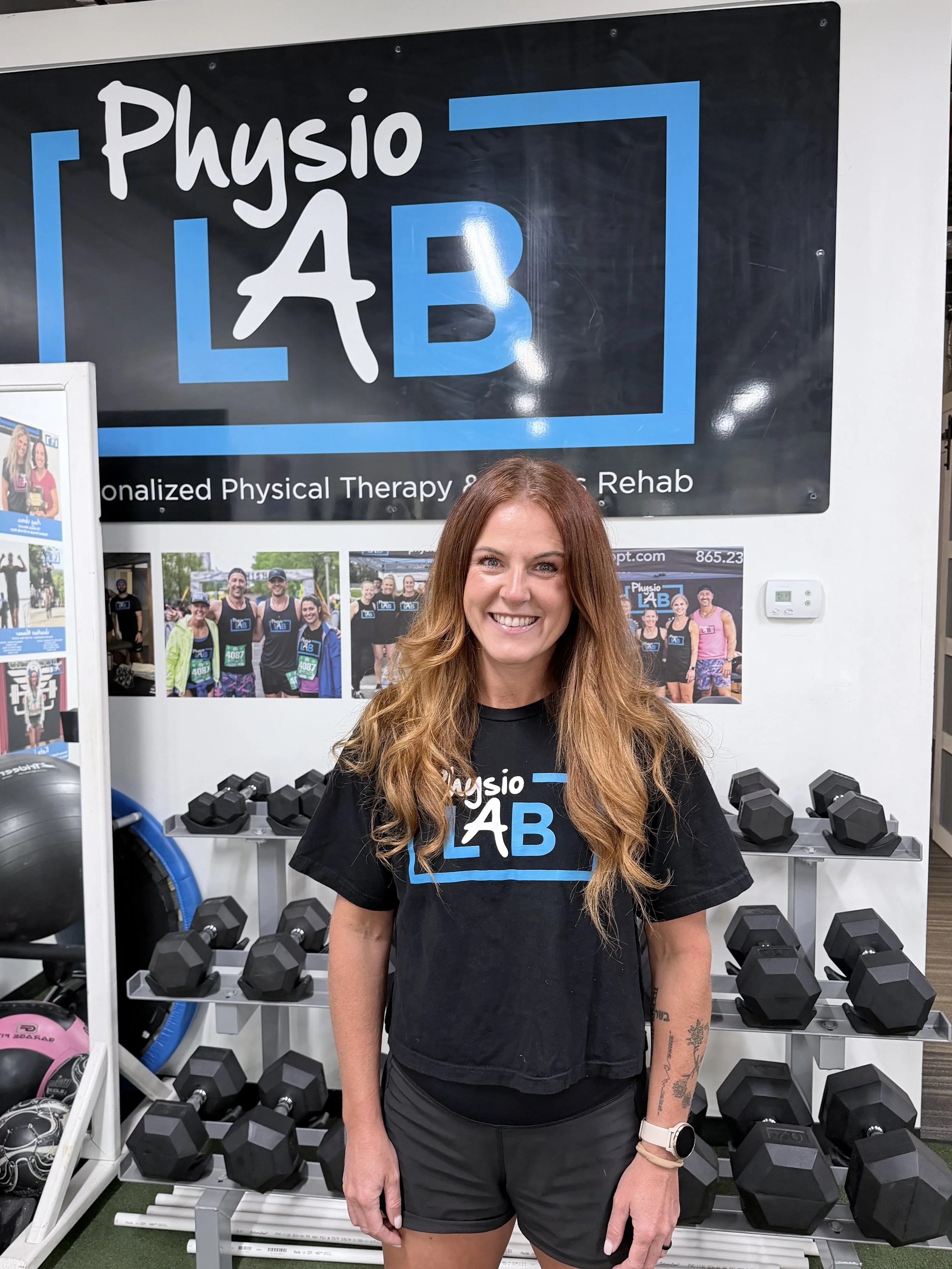 A woman with long reddish hair smiling, standing in front of a display with various dumbbells, at a fitness or physical therapy lab called 'Physio LAB.'