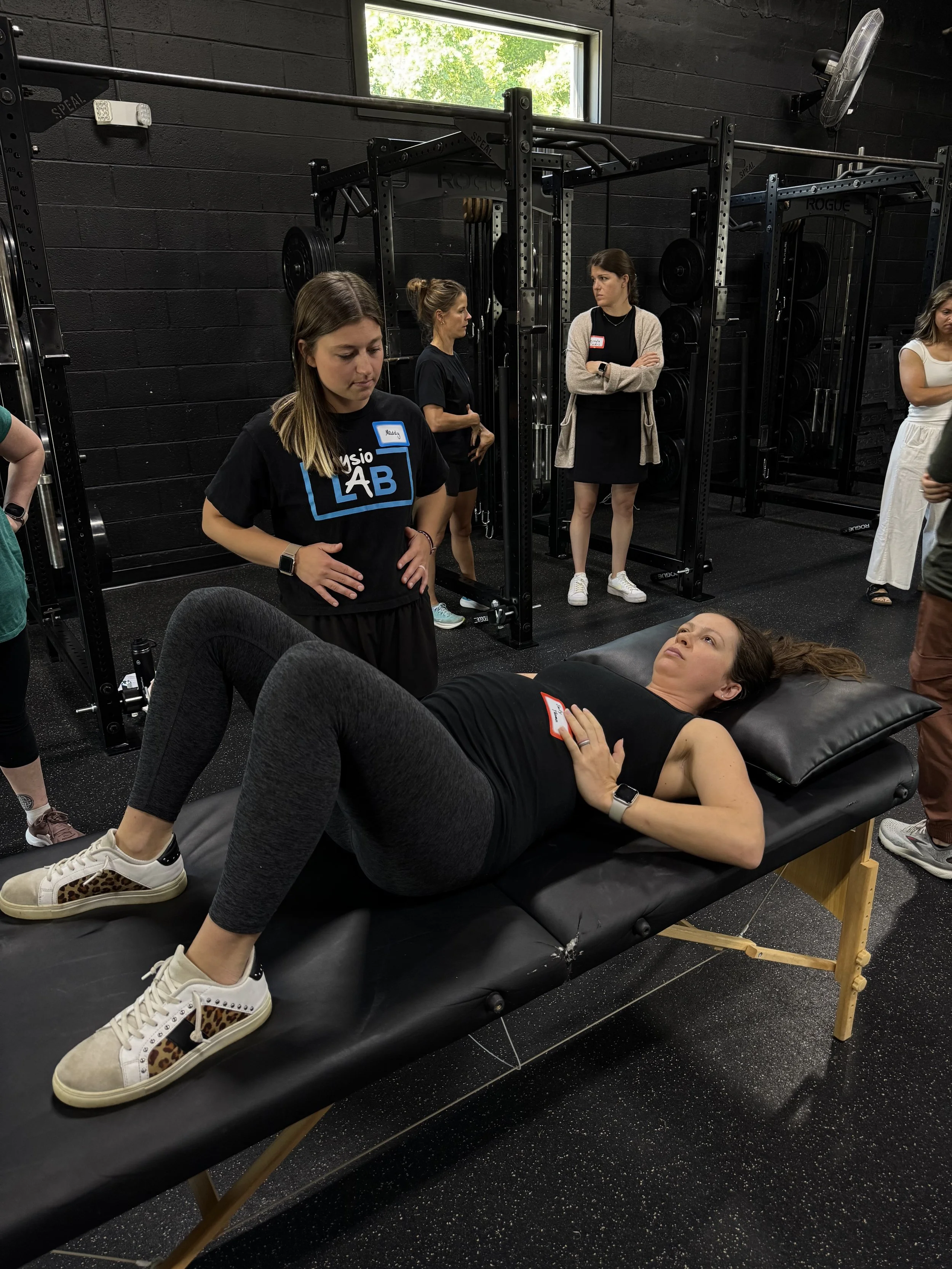 Women in a gym standing and lying on a massage table, with weightlifting equipment in the background.