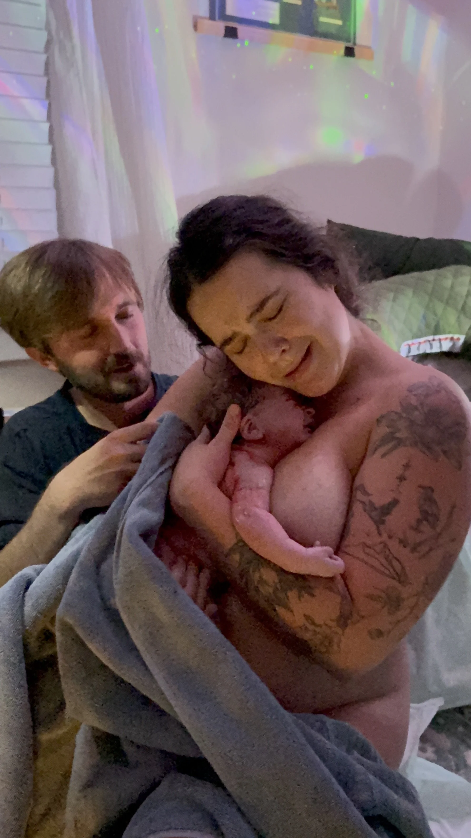 A freshly birthed baby is cradled to their mother's chest. Her partner comforts her in the background.