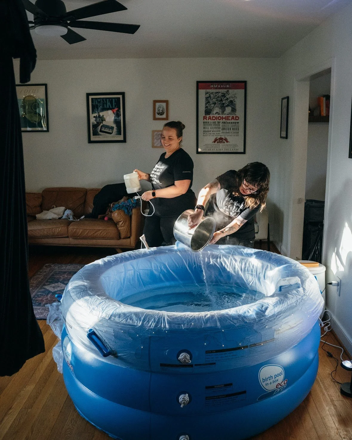 Two Doulas dressed in black shirts and pants pouring hot water from kitchen pots into a large blue inflated birth tub. They are laughing and smiling.