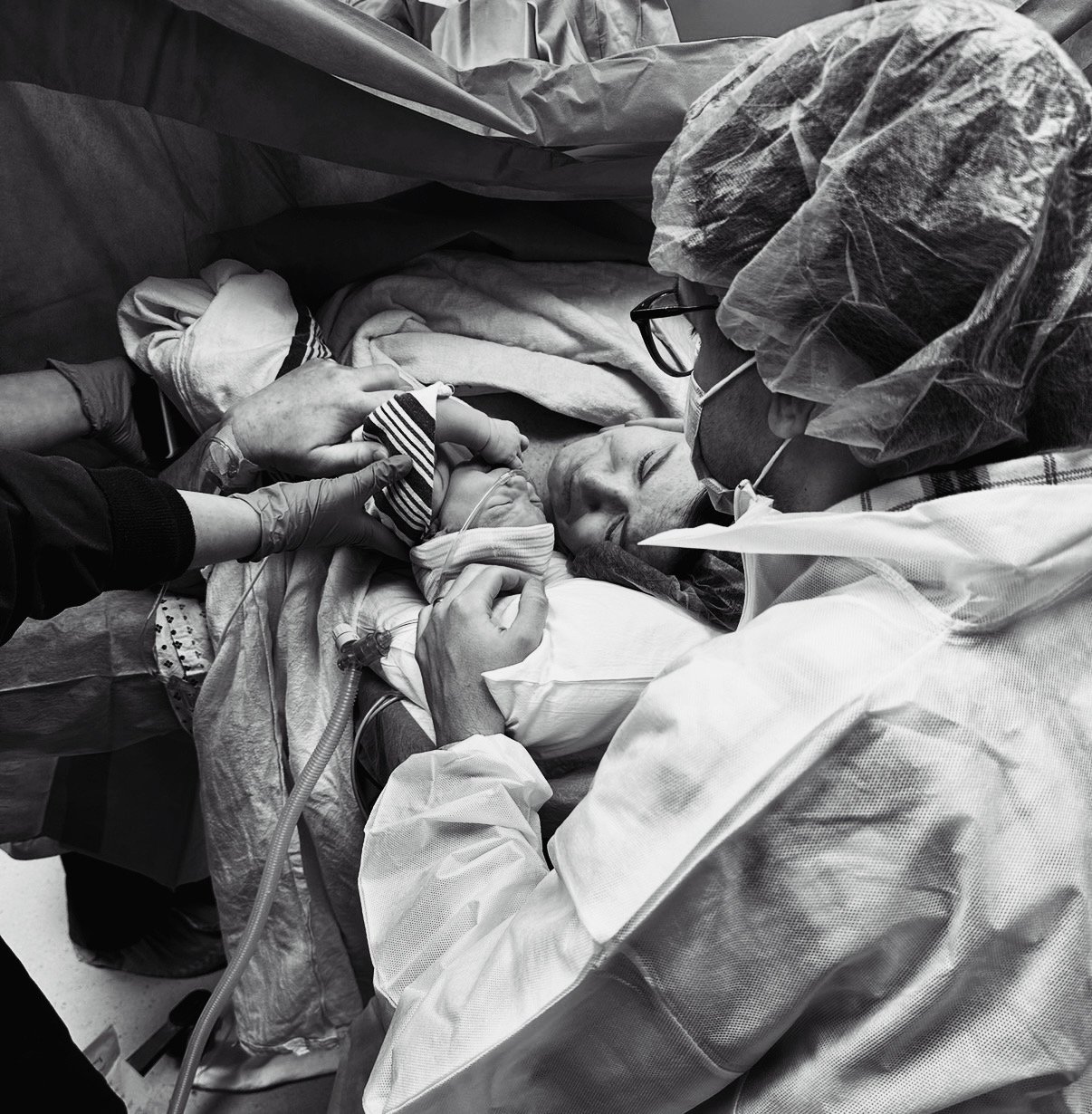 A black and white image of a family during a cesarean birth. a gloved hand holds a swaddled baby to their mother's face.