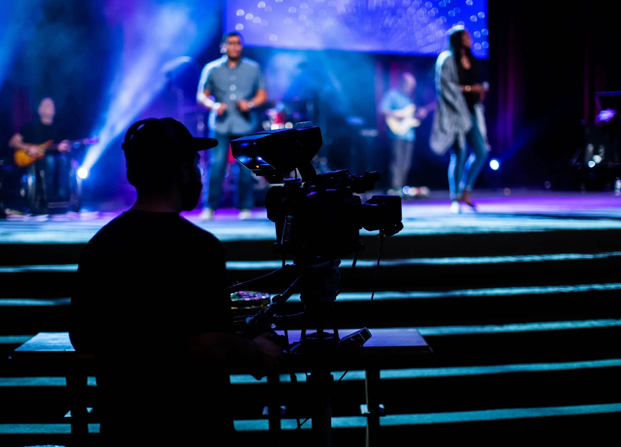 Silhouette of a cameraman filming a live band on stage with colorful lights.