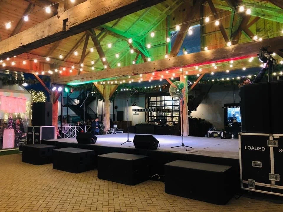 Empty stage with microphones, speakers, and black boxes in a rustic, wooden event space decorated with string lights and green lighting on the ceiling.