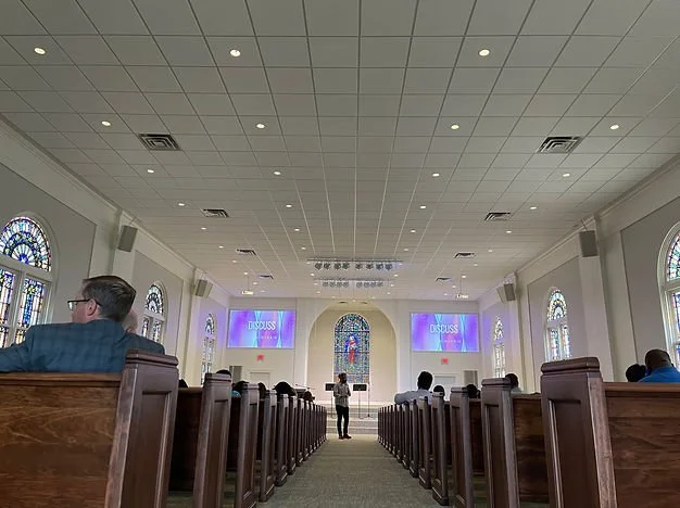 Interior of a church with wooden pews, stained glass windows, and a stage with two screens displaying the word 'DISCUSS' and a person speaking at the front.
