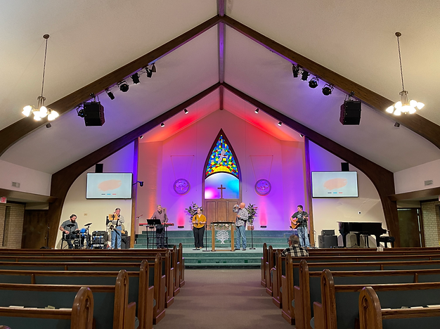A church interior with a worship band performing on stage, stained glass window, and worship leaders singing, with pews in the foreground.