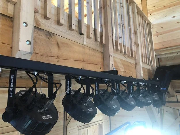 Stage lighting equipment hanging from a black bar attached to a wooden ceiling and railing, in a cabin interior.