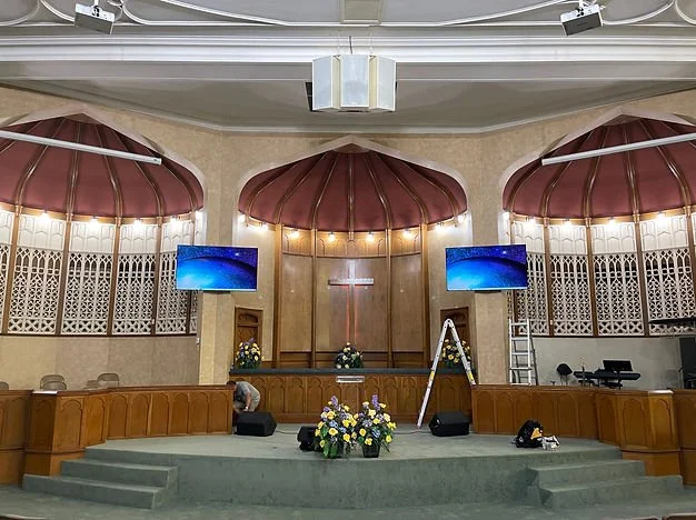 Interior of a church sanctuary with a cross on the wall, two curved wooden railings, and a stage decorated with flower arrangements. Two large display screens hang on either side of the cross, and a ladder is positioned near the stage. The ceiling ha