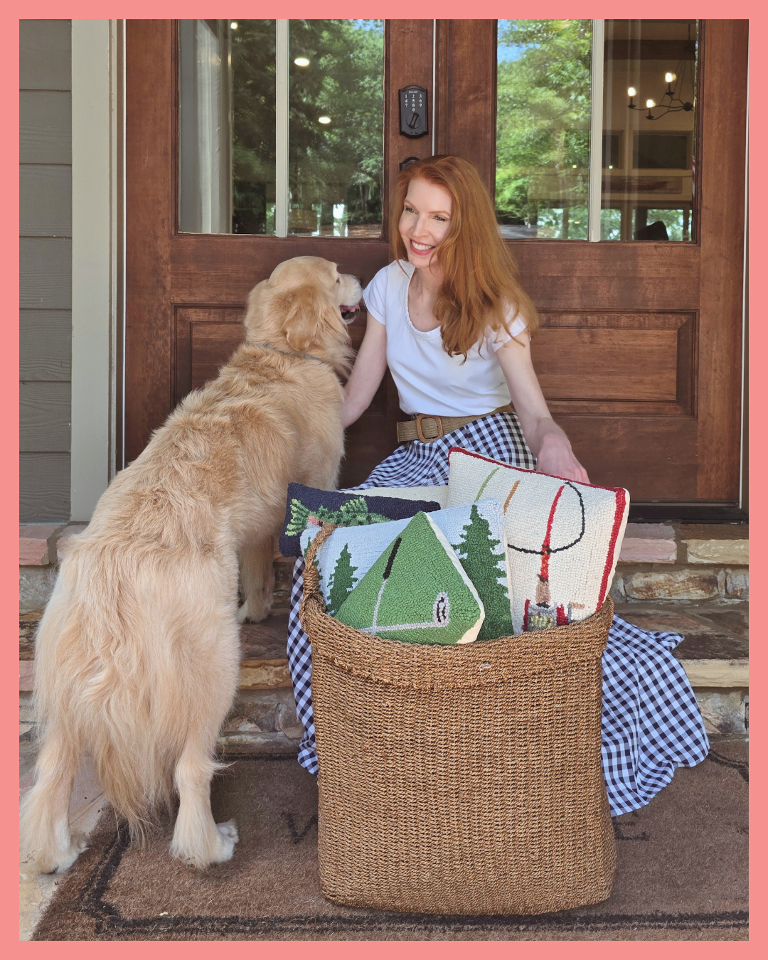 A woman with long red hair sitting on the porch steps in front of a wooden door, smiling at a golden retriever puppy. The puppy is standing on the steps and facing the woman. There is a large woven basket on the ground in front of them, filled with decorative pillows with green trees, a hockey stick, and a snow globe designs. A checkered cloth is draped over the steps.