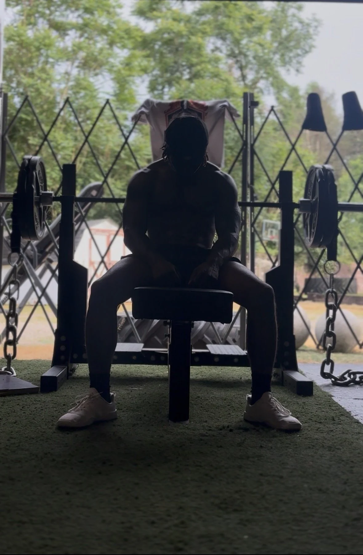 A person sitting on a bench in front of a large window, with trees visible outside. The person's face is obscured by shadows, and they are wearing athletic clothing and sneakers. There is gym equipment around them, including a barbell and chains.