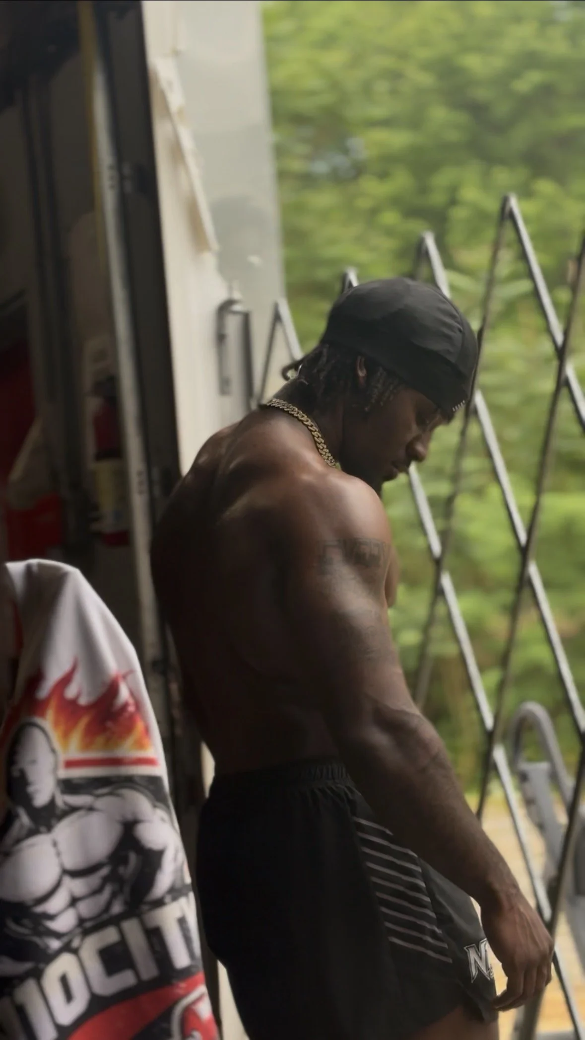A shirtless man with tattoos, wearing a black cap and gold chain, standing near a window with green trees outside, with a partially visible skateboard and a person wearing a hoodie in the foreground.