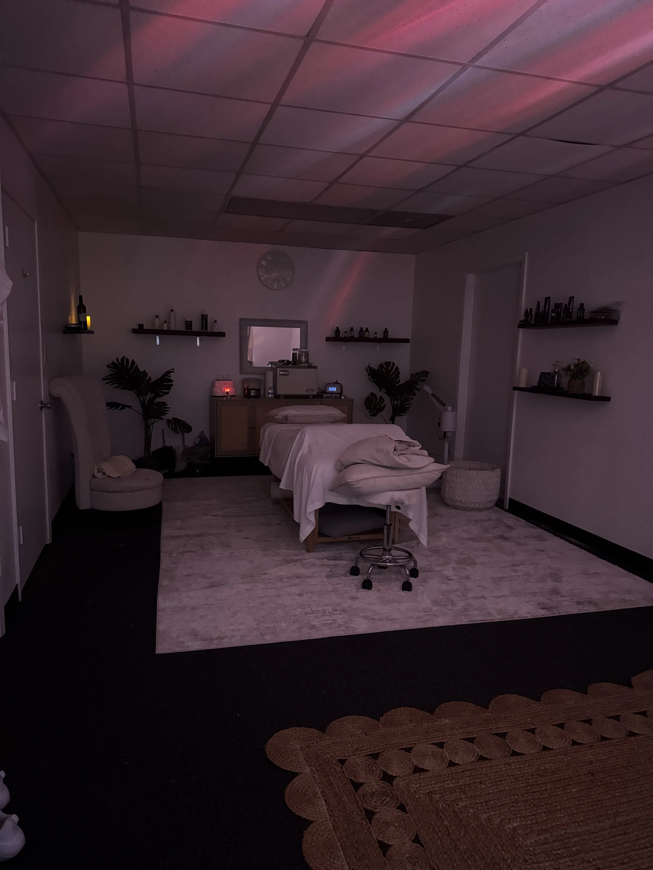 A dimly lit massage therapy room with a massage table covered in white sheets, a rolling stool, two plants, wall-mounted shelves with bottles, a clock, and various therapy equipment, illuminated by subtle pink and purple lighting.