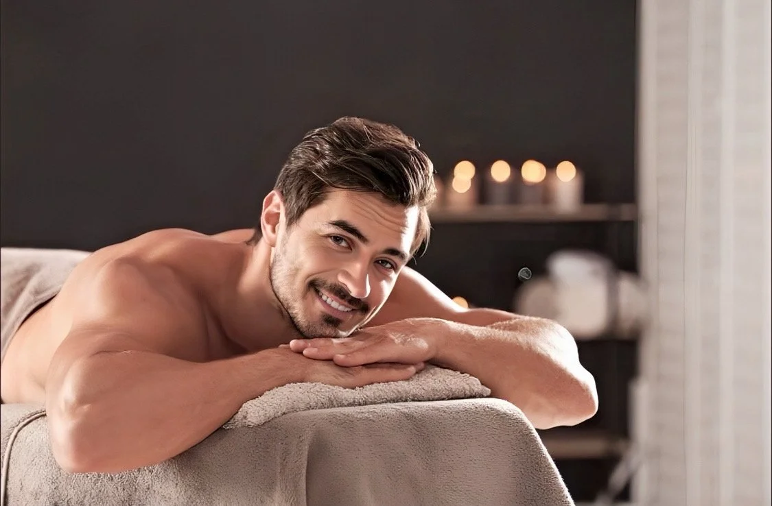 A man lying face down on a massage table, smiling at the camera, in a cozy room with candles and soft lighting.