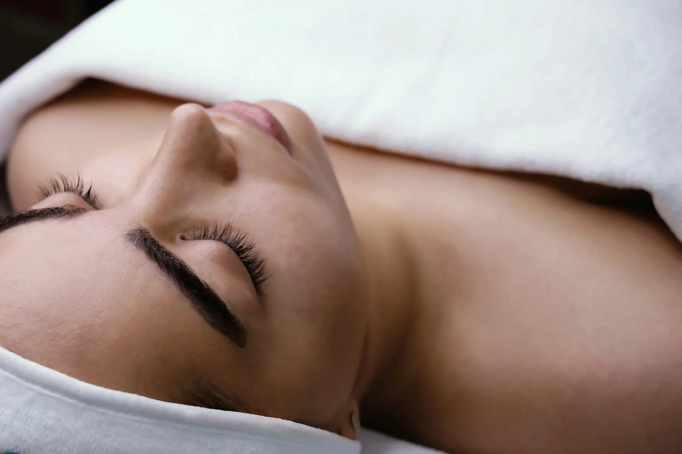 Close-up of a woman with long eyelashes, his eyes closed, lying on her side with a white cloth partially covering her face and head.