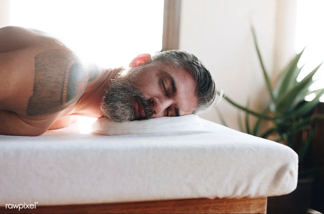 A man with a beard and tattoos sleeping on his side on a mattress with a white pillow in a room with natural light and a plant in the background.