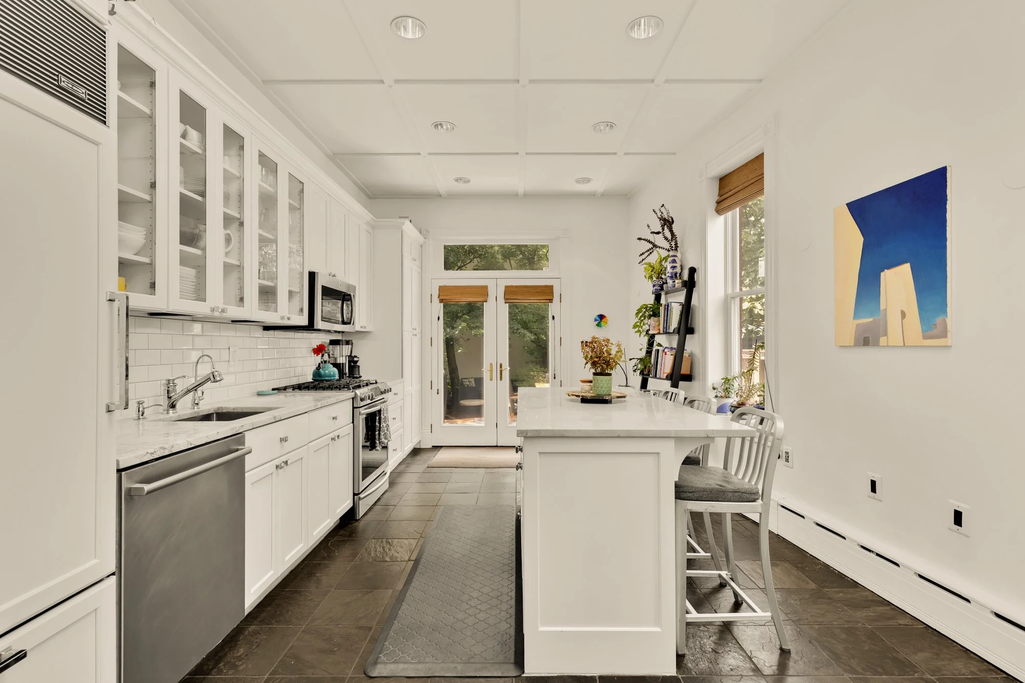 A white kitchen with white cabinets, a marble countertop, a stainless steel dishwasher, a stove, a microwave, and a sink. There is a kitchen island with chairs, a gray rug, and a painting on the wall. The room has windows with wooden blinds and a doo