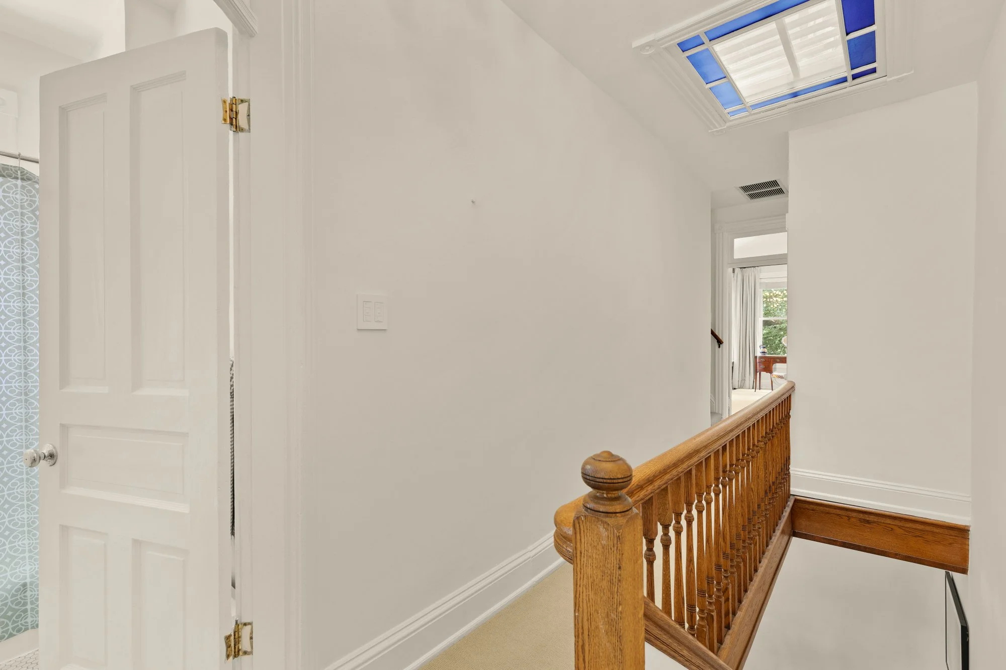 View of a staircase landing area with white walls, a wooden railing, a skylight window, and a door on the left that is slightly open, revealing part of a patterned curtain.