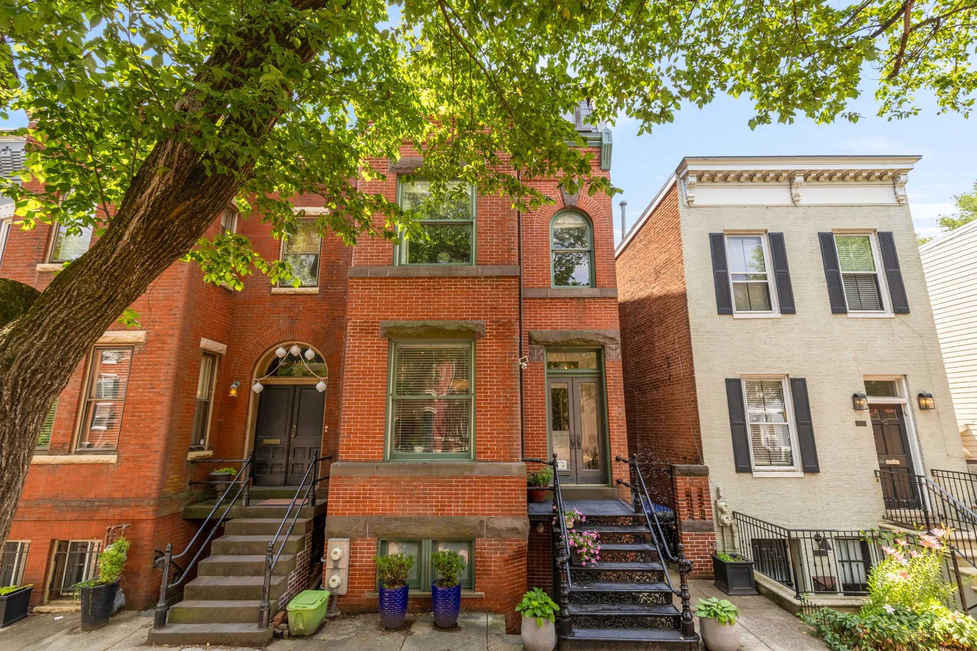 Three urban row houses with brick facades, stairways, and entering doors, set against a bright sky with tree shade.