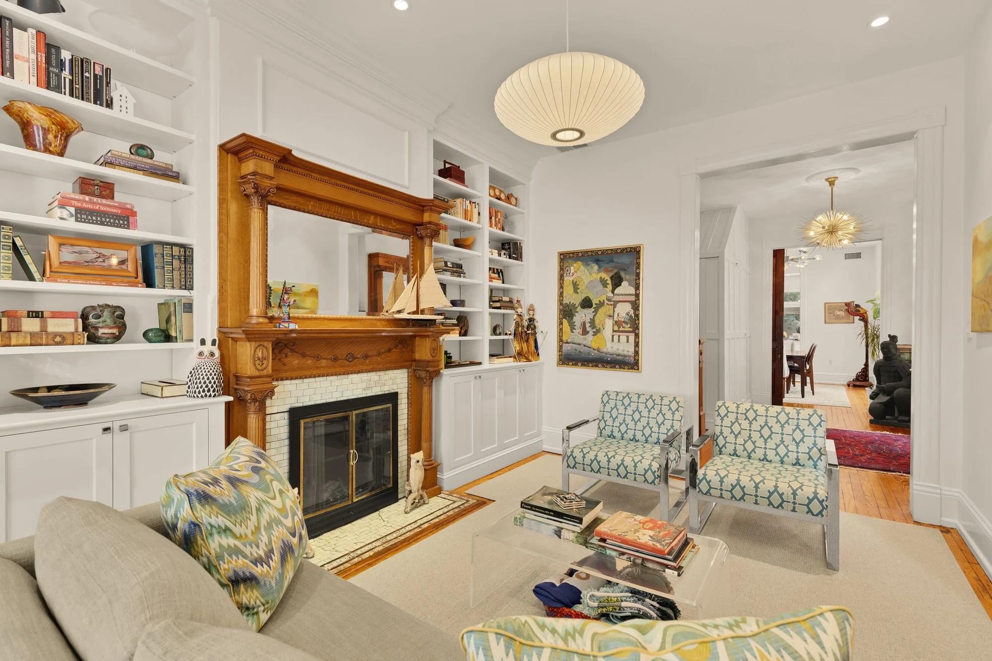 Living room with a white-painted wood mantel and built-in bookshelves, decorated with books and art. In front is a beige sofa with colorful pillows, a glass coffee table with books, and patterned armchairs. There are two ceiling lights, one drum-shap