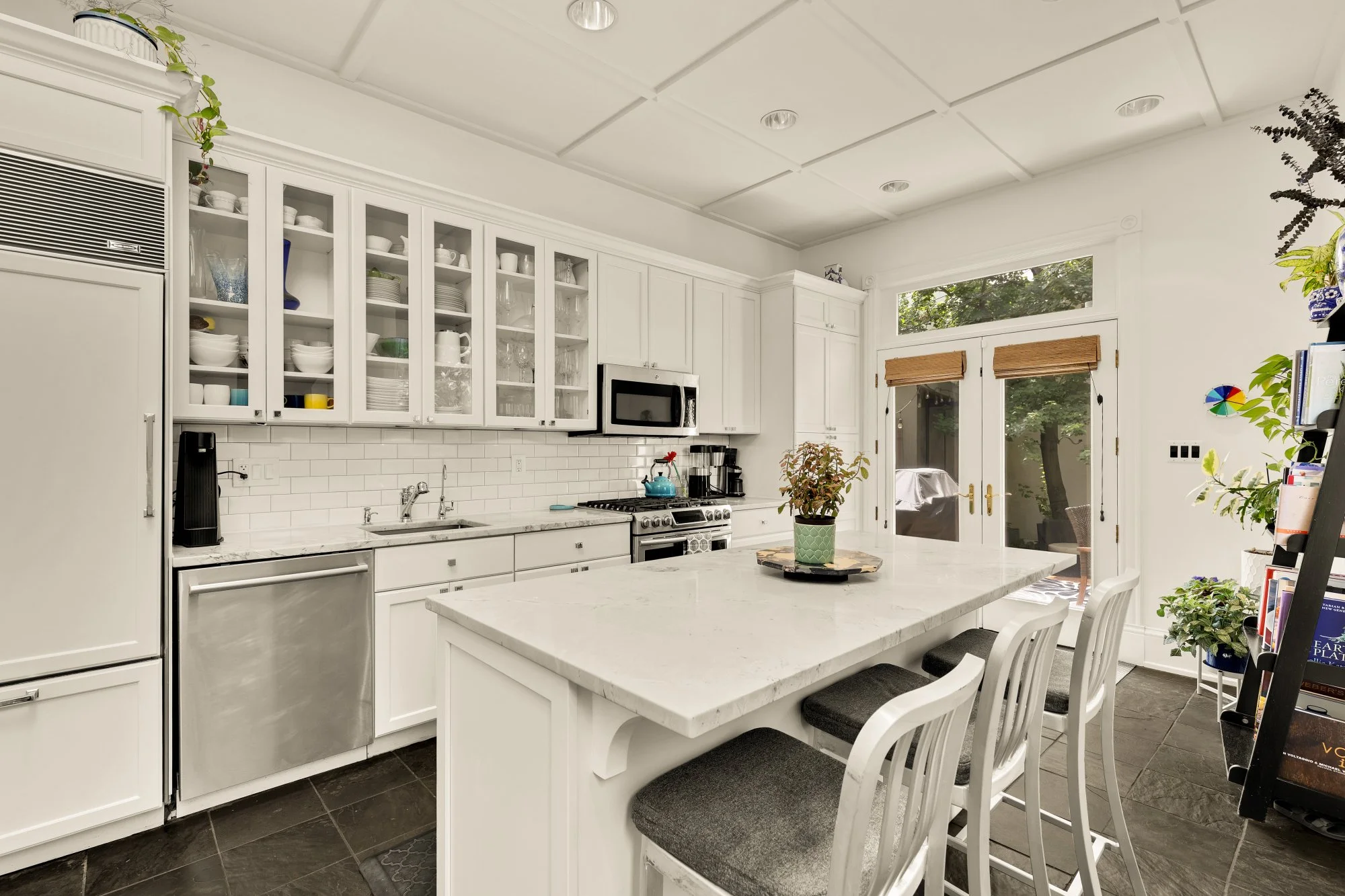 A bright, white kitchen with open shelving cabinets filled with dishes, glasses, and bowls. There is a stainless steel refrigerator, a microwave above the stove, and a countertop with a coffee maker. A large central island has a potted plant, and a d