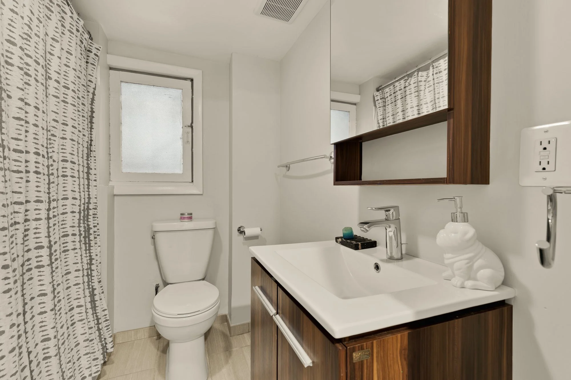 A small bathroom with a white toilet, a wooden vanity with a white sink, and a mirror. There is a patterned shower curtain on the left, a frosted window above the toilet, and a soap bottle shaped like a bear on the sink. The walls are painted white.