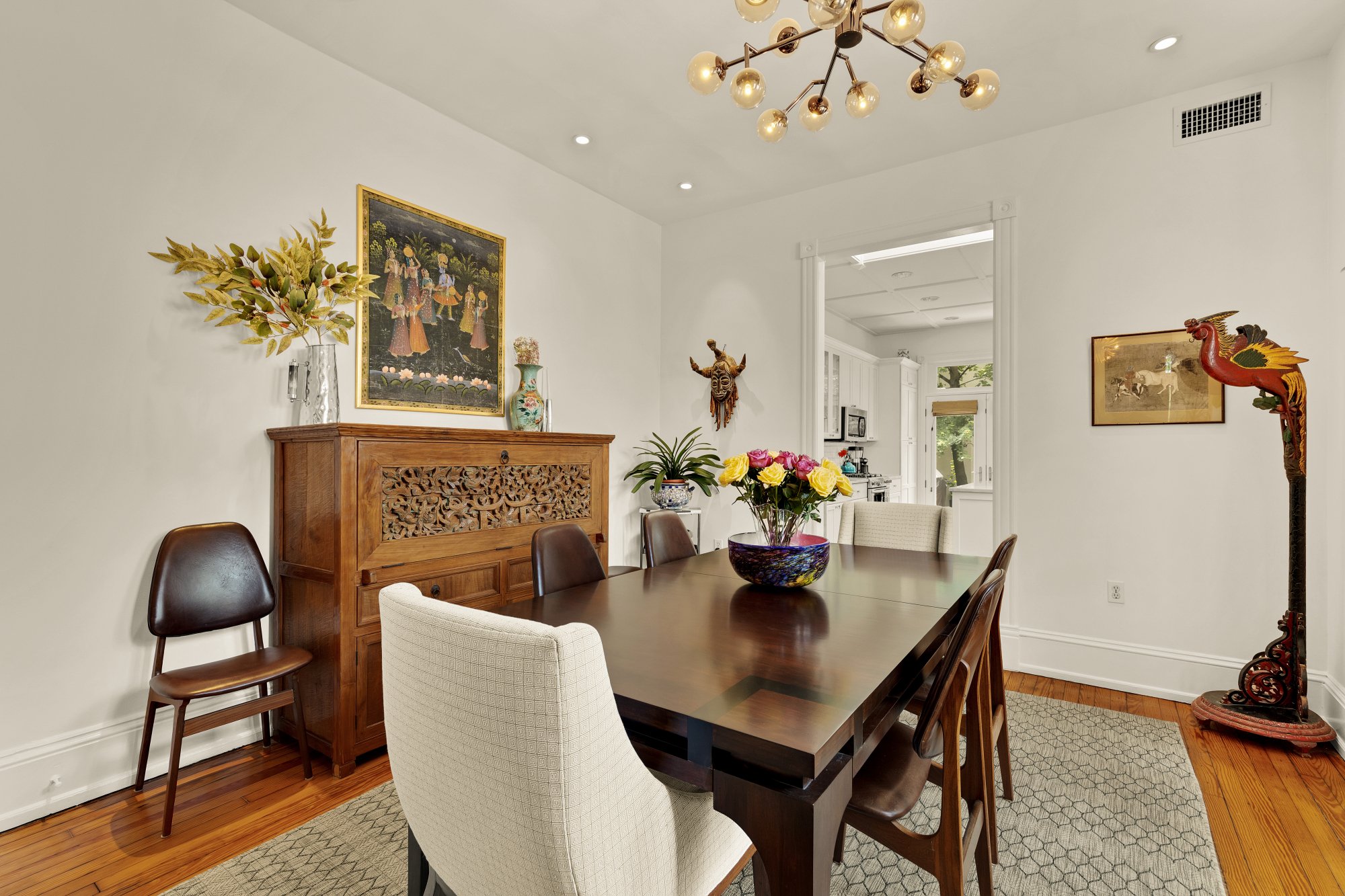 A dining room with a dark wood table surrounded by six chairs, a large colorful floral vase on the table, a wooden cabinet against the white wall with artworks and decorative items, a modern chandelier, and a doorway leading to a kitchen with a window and white cabinets.