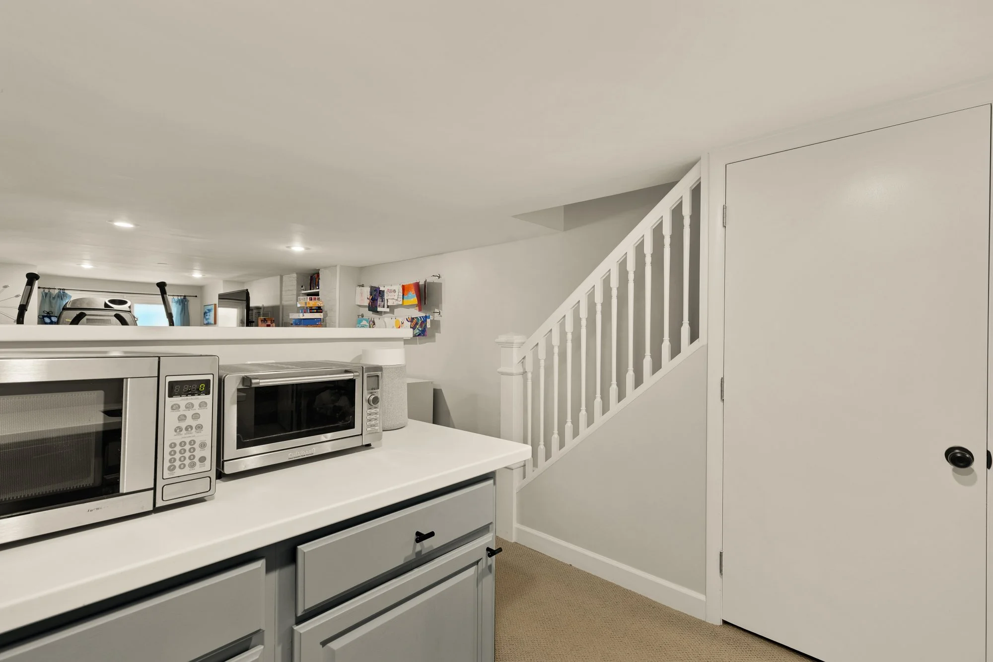 A kitchen with a white countertop, a Microwave oven, a toaster oven, and a small speaker. In the background, there is a staircase with white balusters, a door, and a wall shelf with children's drawings and books. The kitchen has a white ceiling with 