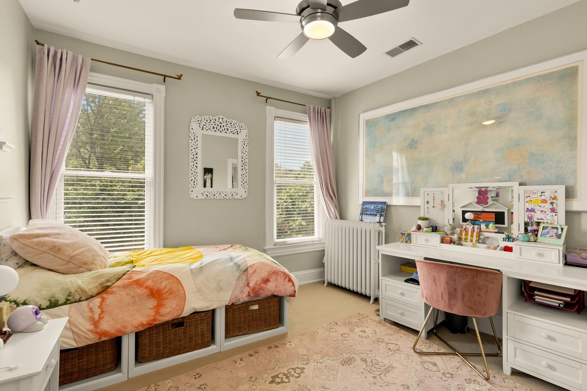 A cozy bedroom with a bed, a window with pink curtains, a decorative mirror on the wall, a ceiling fan, a desk with jewelry storage, framed artwork, and a pink velvet chair. The room has light green walls, beige carpet, and a patterned area rug.