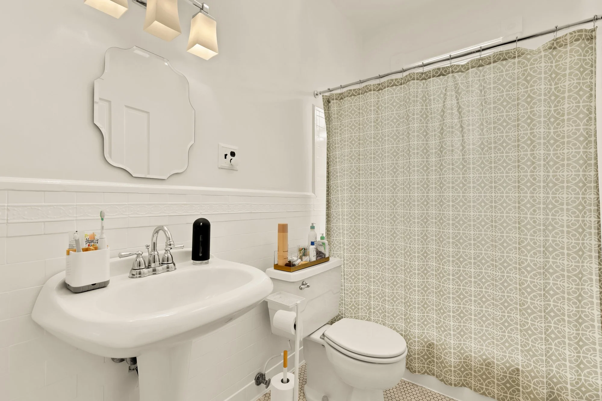 A bathroom featuring a white sink with toiletries, a mirror, and a window with patterned curtain, next to a toilet with a roll of toilet paper, and a cleaning brush.