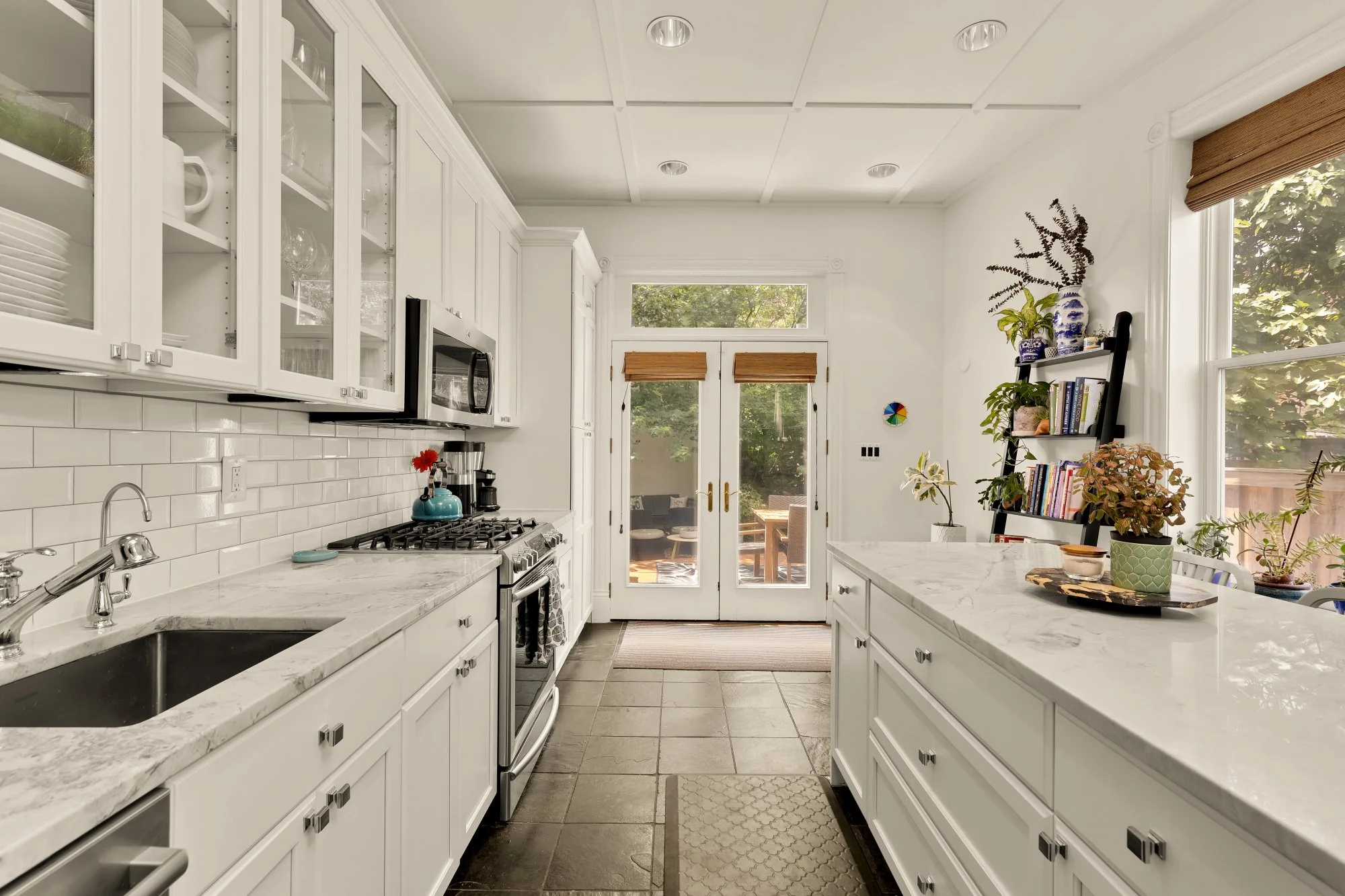 Kitchen with white cabinets, marble counters, window and door to outside patio, plants, and a bookshelf.