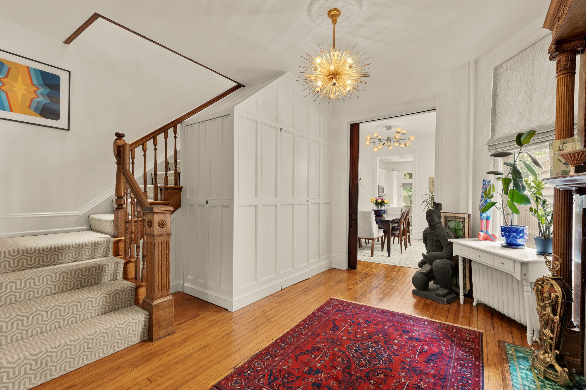 A spacious entryway with wooden stairs, a colorful abstract framed painting on the wall, a bright chandelier, a red patterned rug, potted plants, and decorative sculptures.