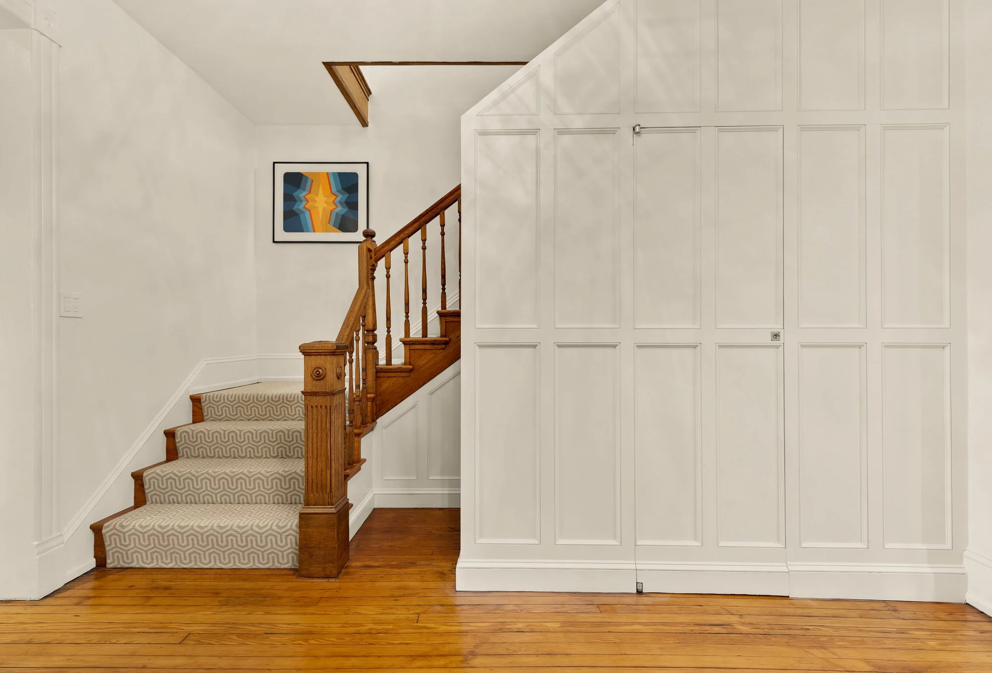  Interior of a house with a staircase, framed abstract art, white paneled wall, and hardwood flooring.