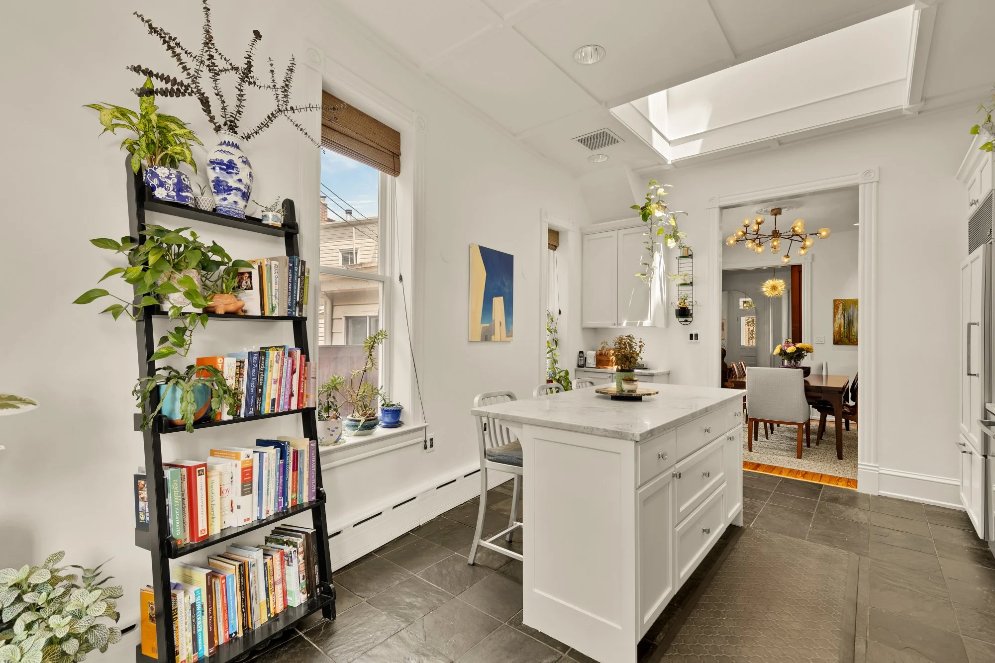 A bright and airy kitchen with a white kitchen island, white walls, and a white ceiling with skylights. There are plants and decorative items on a bookshelf and windowsill, and a small dining area with chairs and a table in the background. The space 