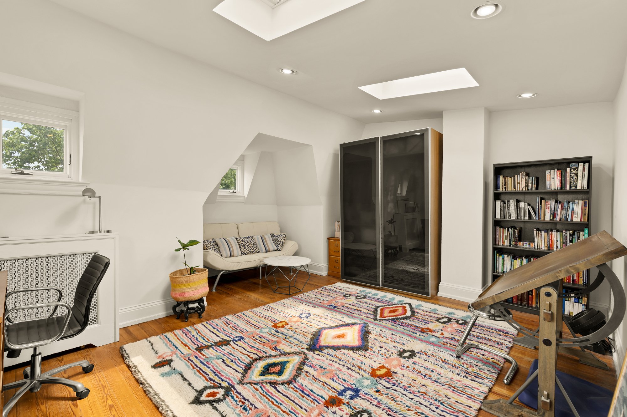 Cozy living room with white walls, hardwood floors, a vibrant multicolored area rug, a white sofa with patterned pillows, a small white round coffee table, bookshelves filled with books, small windows, a black and gold wardrobe, and a wooden dining t