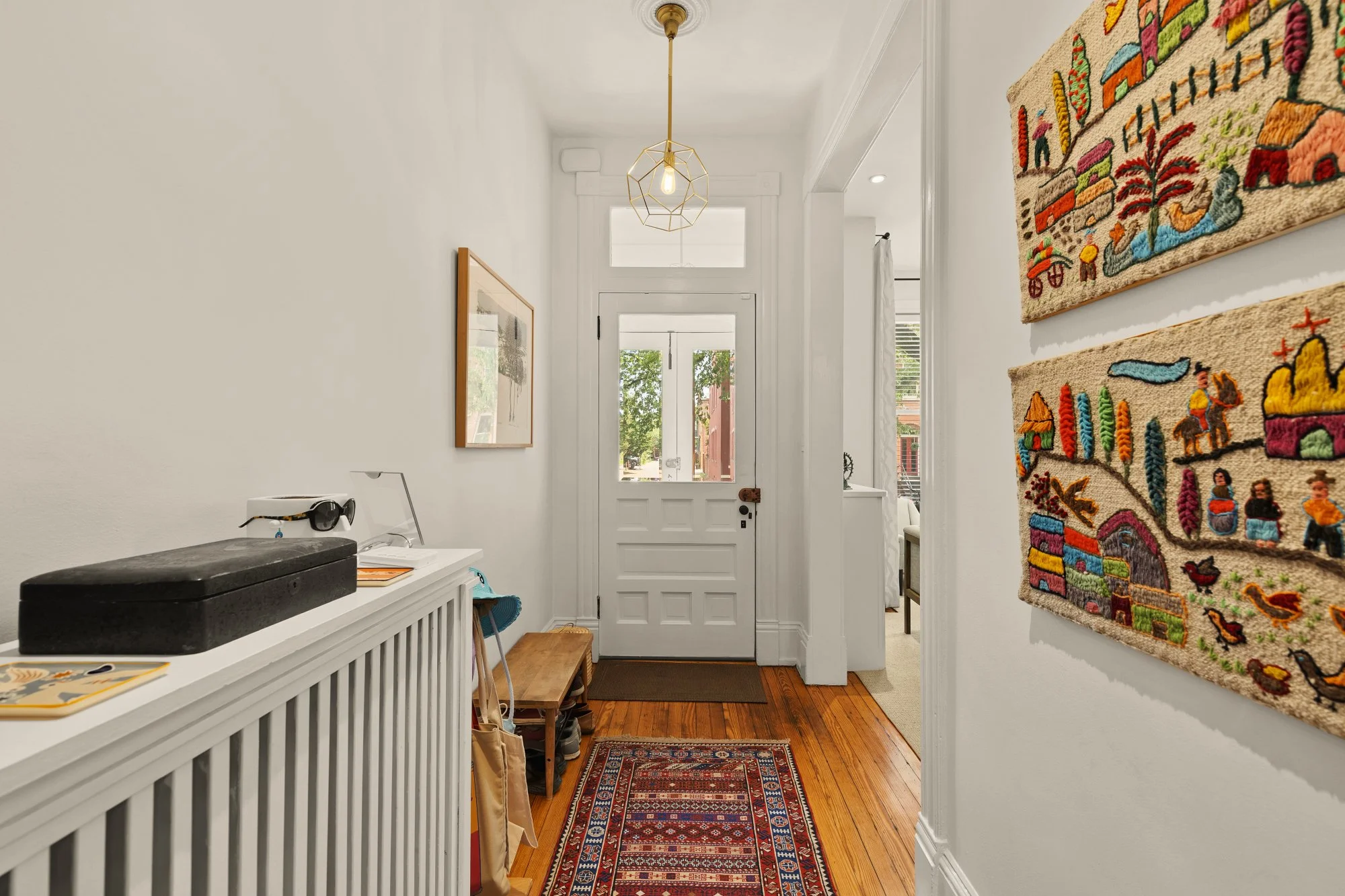 A hallway with white walls and hardwood floor, decorated with colorful woven textile art and a framed picture, leading to a door with a window showing trees outside. There is a black printer on a white radiator cover, a small bench, and a patterned r