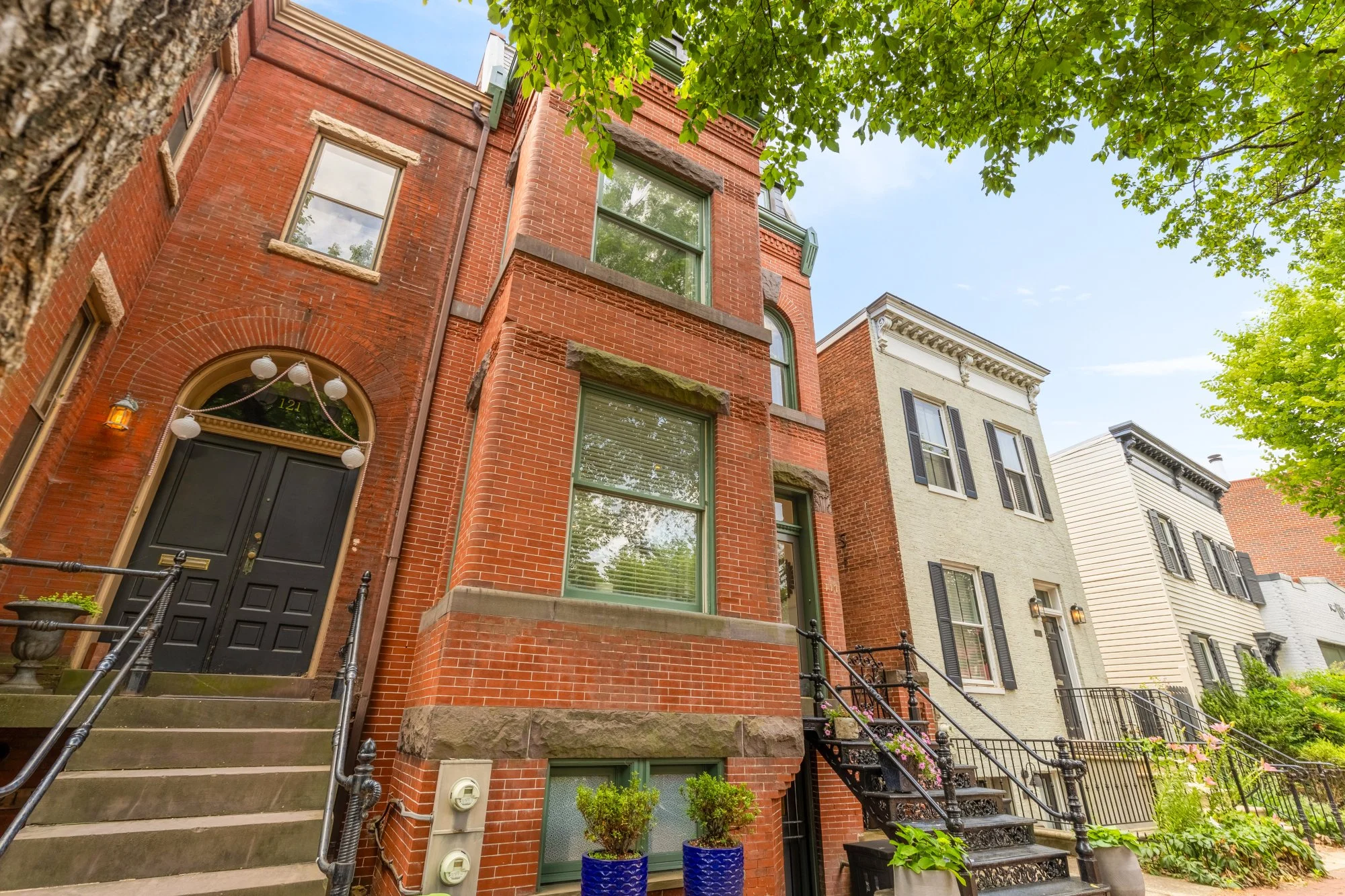 Street view of brick townhouse with black door, stairs, and potted plants in blue containers, framed by green trees and neighboring homes.
